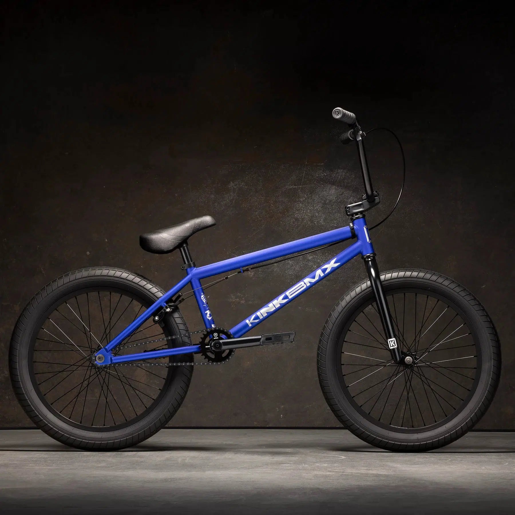 The Kink Curb 20 Inch Bike, featuring blue color, Mission Tracker Tyres, and Kink Ace Grips on its handlebars, stands out against a dark background.