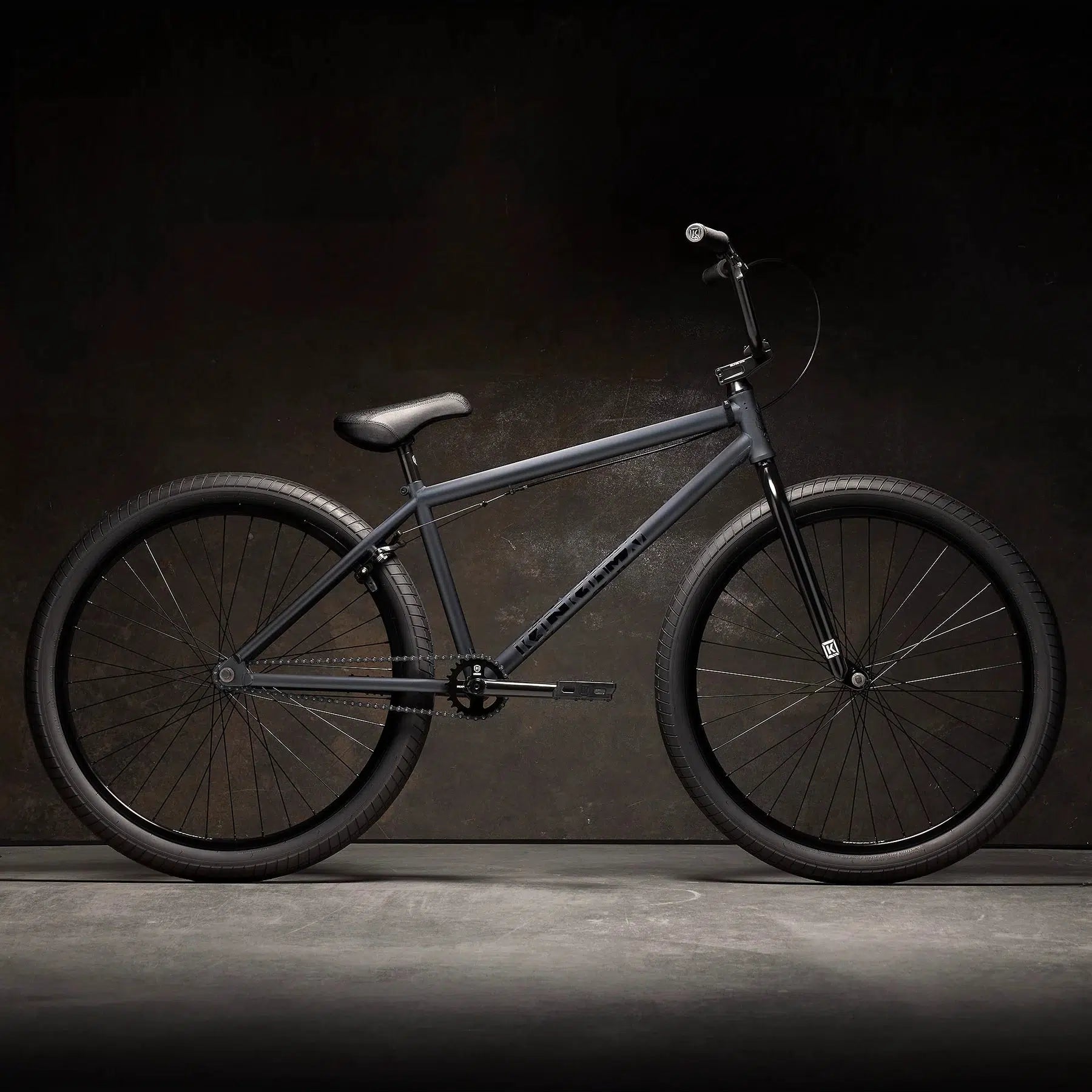 A Kink Drifter 26 Inch Bike in matte black features thick tires, a straight top tube, and a padded seat, set against a dark background. This versatile BMX-style bike is perfect for riders of all levels.