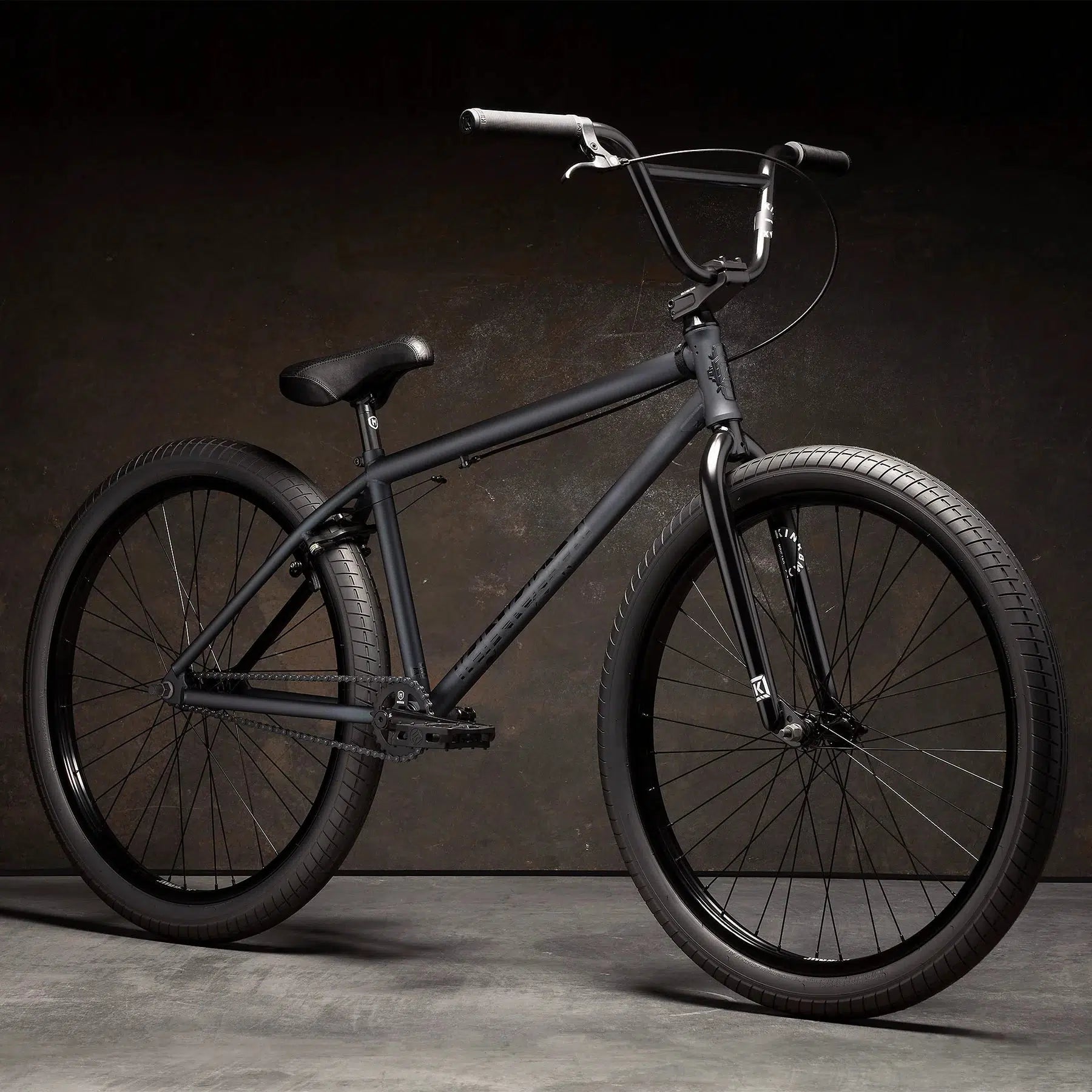 A Kink Drifter 26 Inch Bike in matte black, featuring wide tires, a high handlebar, and a padded seat, positioned against a dark background. This bicycle exemplifies versatility in both form and function.