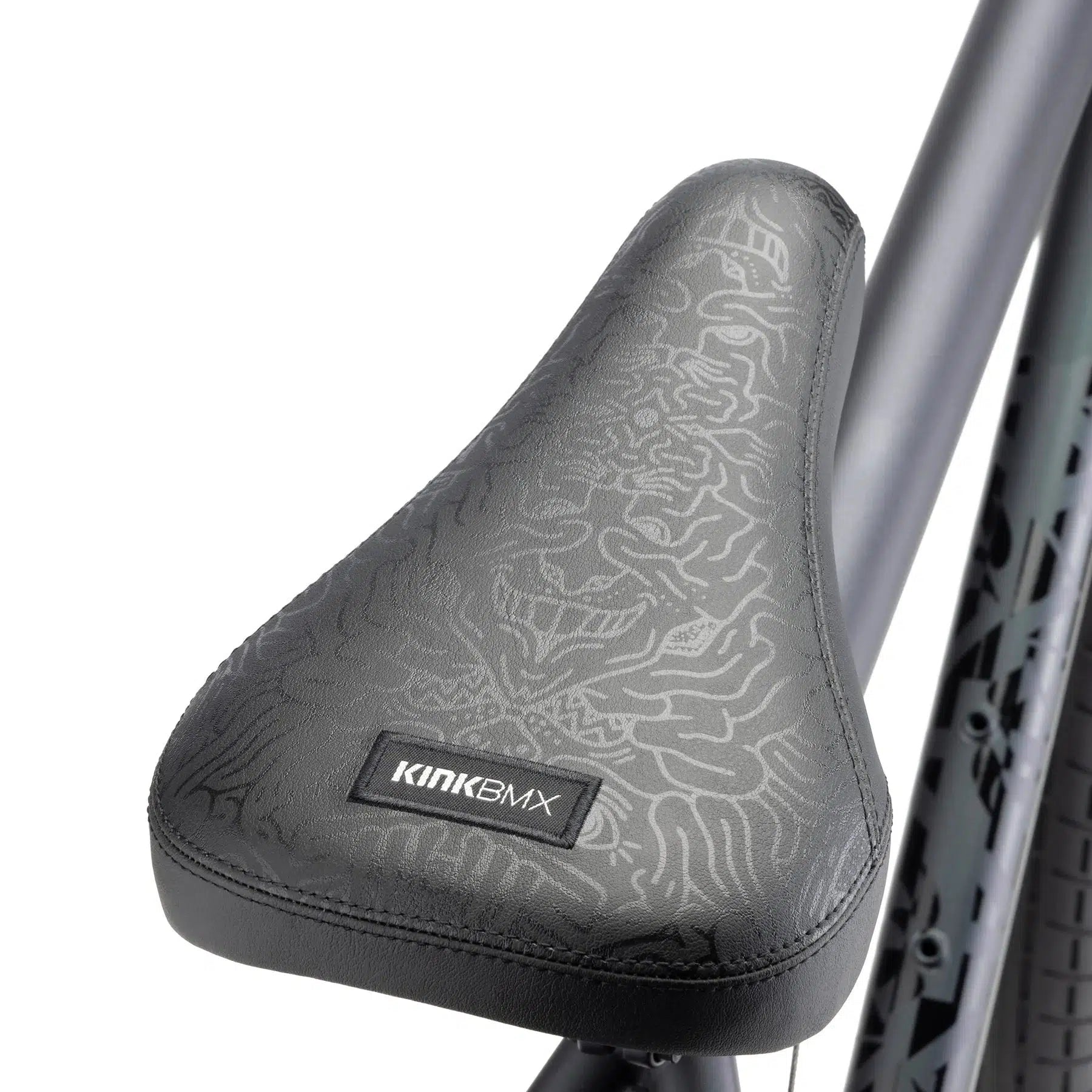 Close-up of the Kink Drifter 26 Inch Bike seat with textured black material and the "Kink BMX" label. The bike frame and partial rear wheel of this versatile 26-inch bicycle are visible in the background.