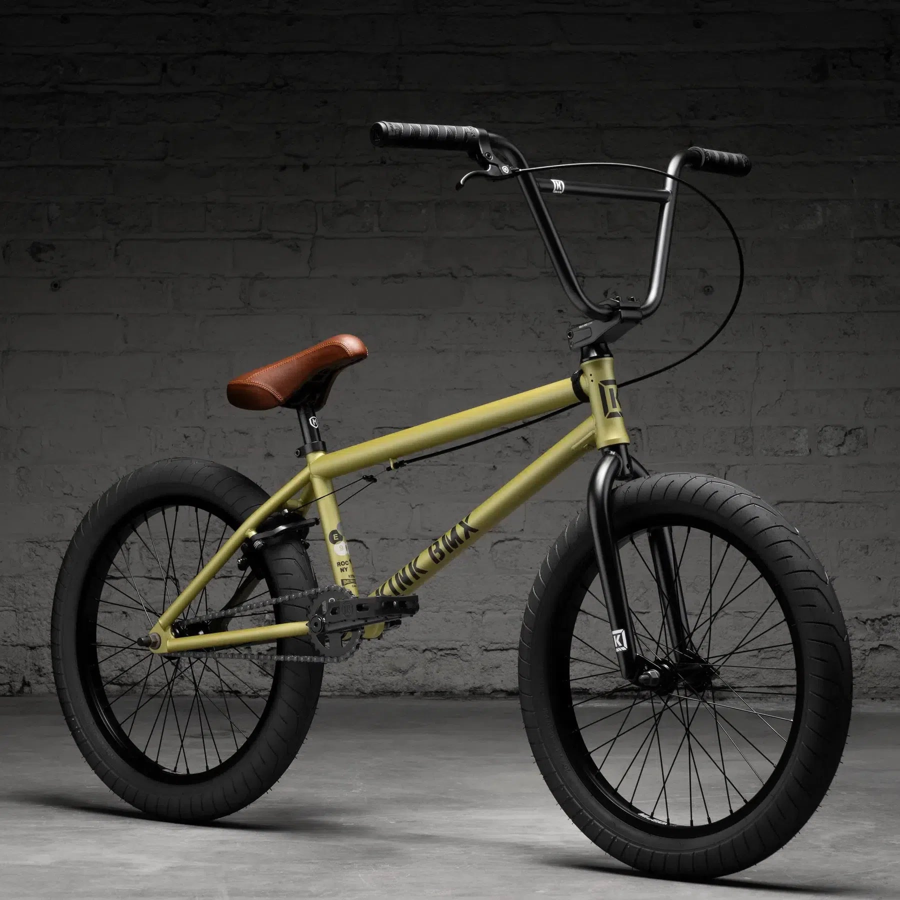 The Kink Gap 20 Inch Bike (2026), featuring a yellow chromoly steel frame with black handlebars and wheels, rests on a gray concrete floor against a brick wall, while its brown saddle offers a striking contrast.