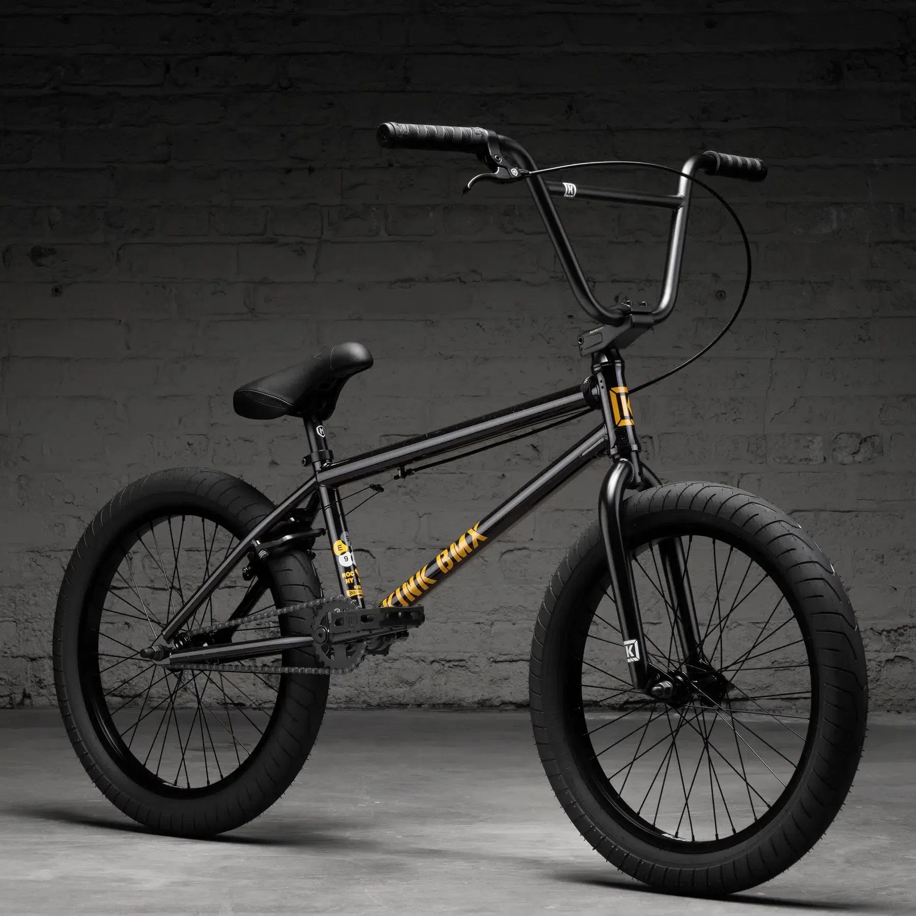 The Kink Gap 20 Inch Bike (2026), crafted from durable chromoly steel, features a black frame with thick tires and yellow decals, set against a gray brick wall.
