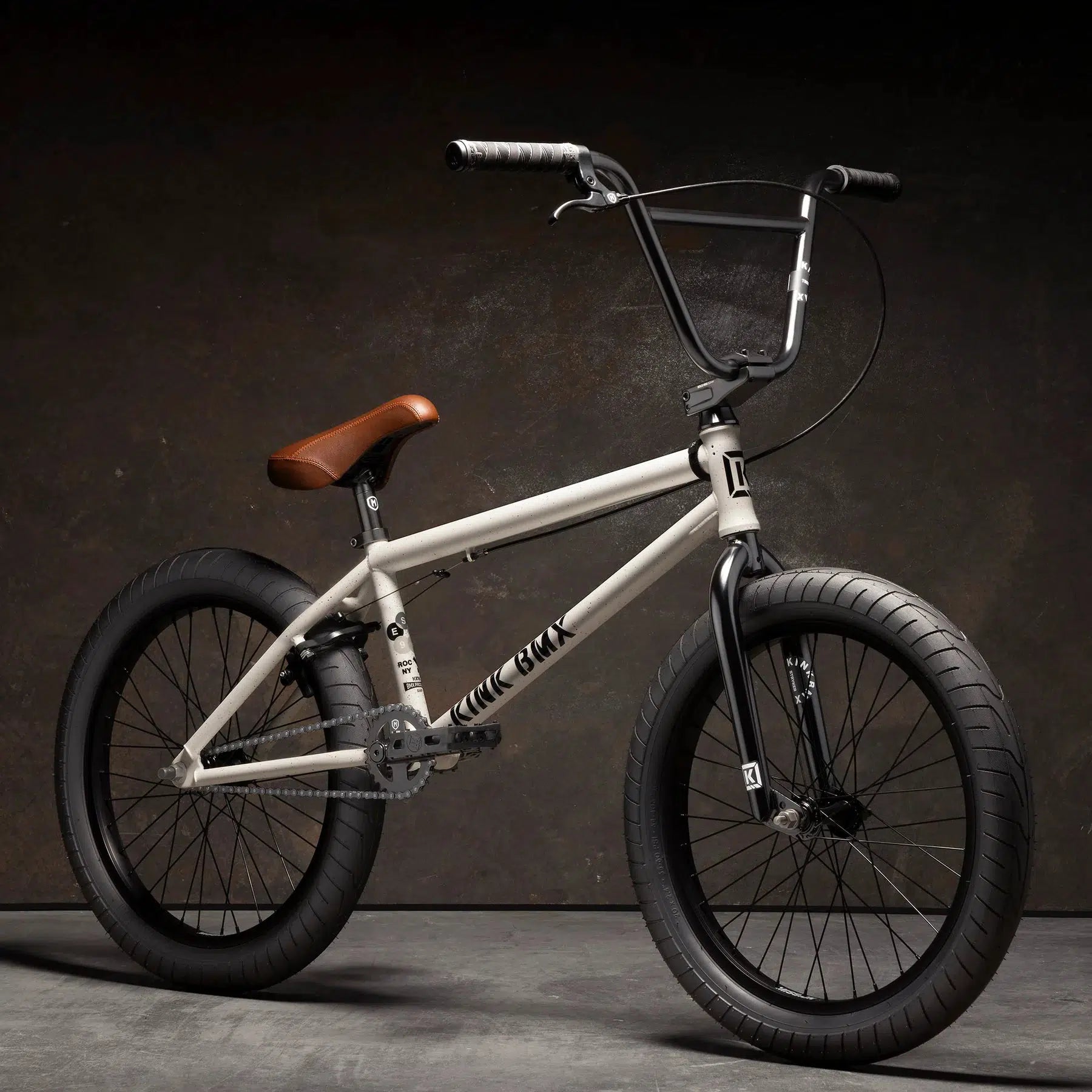 The Kink Gap 20 Inch Bike, with its semi-chromoly light gray frame, brown seat, black handlebars, and thick black tires, is displayed against a dark background.