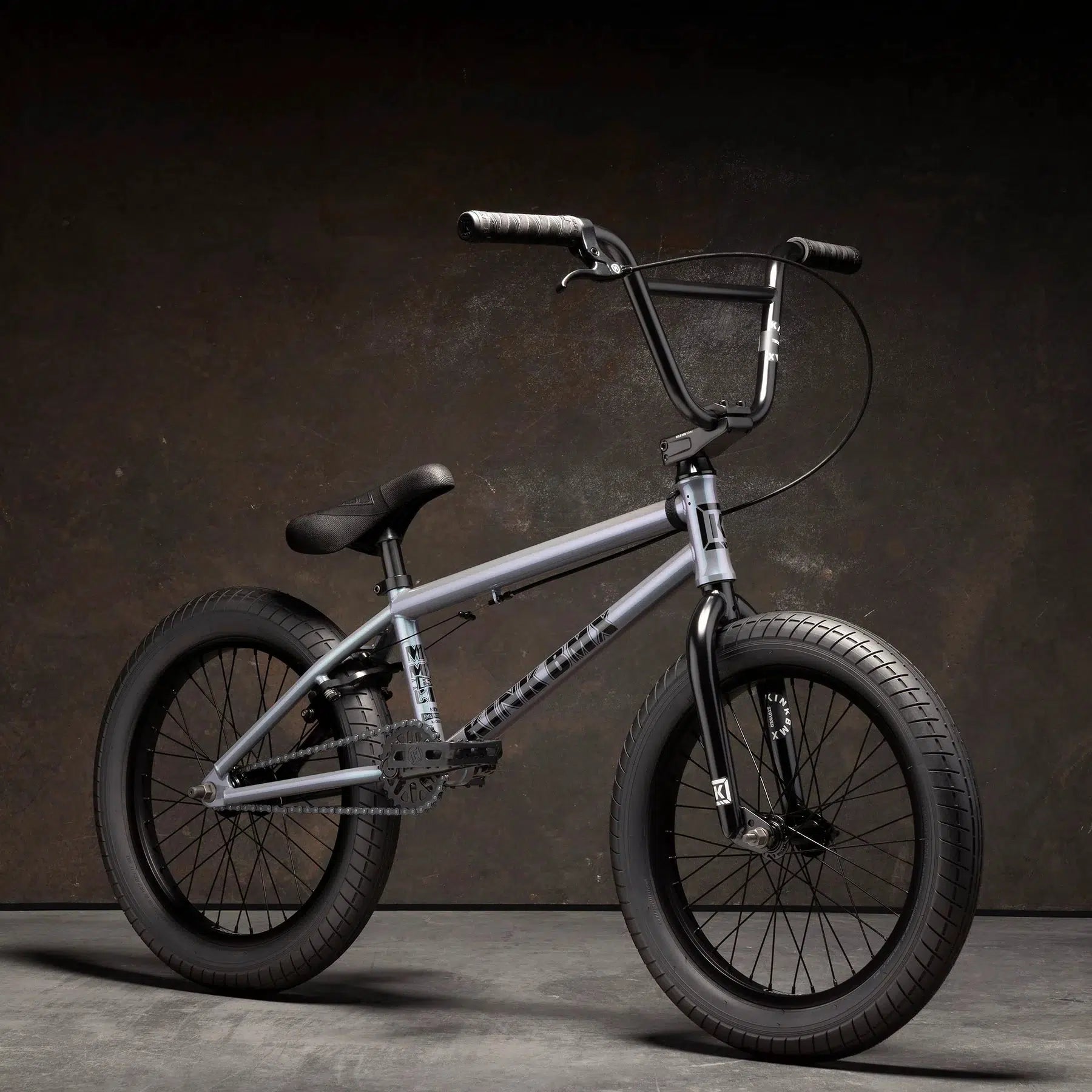 A Kink Kicker 18 Inch Bike with a matte gray frame, thick black tires, and a high-rise handlebar stands against a dark brown textured background. Designed for younger riders, it boasts high-quality components for an exceptional riding experience.