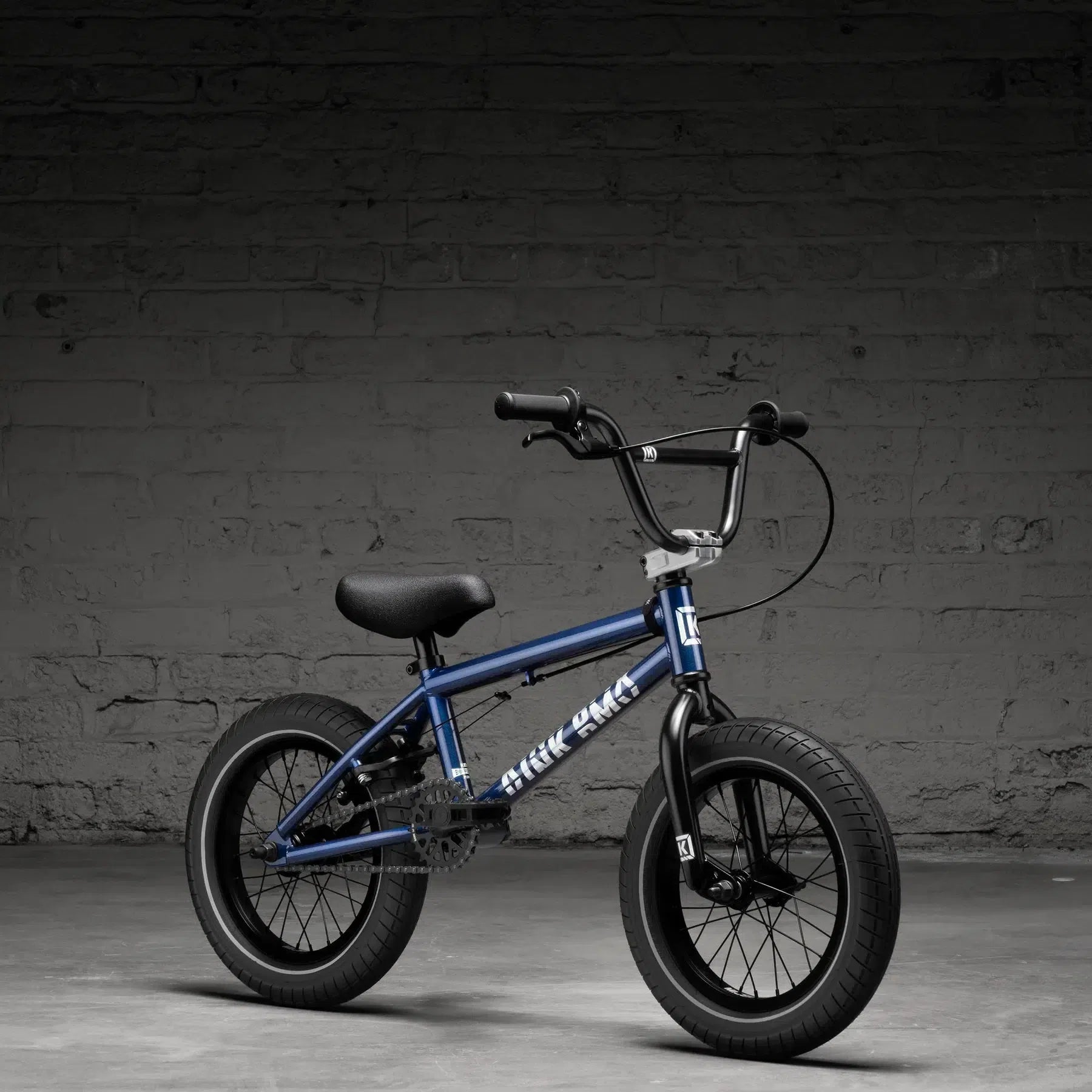 The Kink Pump 14 Inch Bike (2026), featuring a striking blue frame and thick black tires, is displayed against a textured gray brick wall on a smooth concrete floor—ideal for young adventurers seeking thrills.