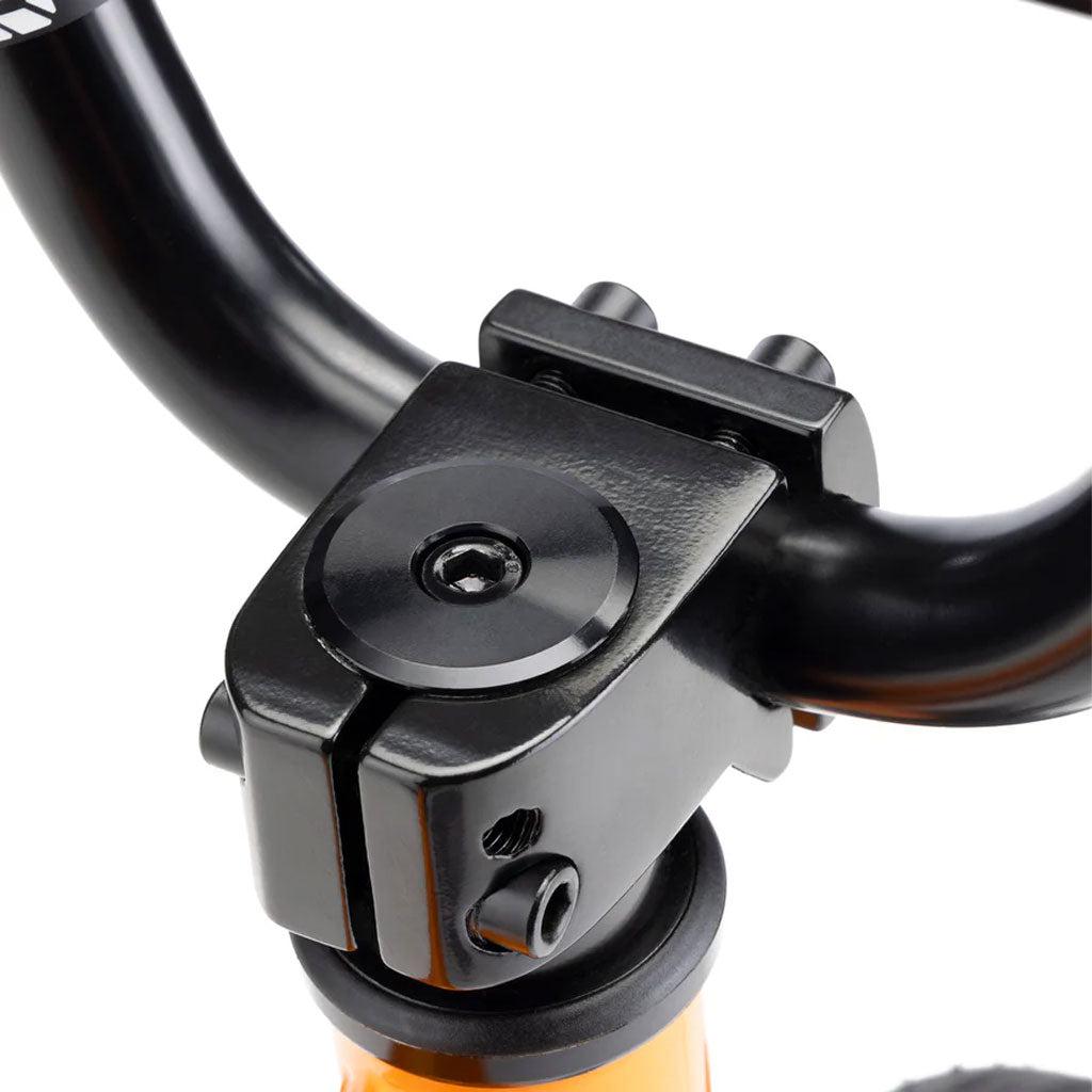 Close-up view of a black bicycle handlebar stem and clamp assembly attached to an orange frame, showcasing the sturdy design of a Kink Roaster 12 inch Bike (2025) with 12” wheels and coaster brakes.