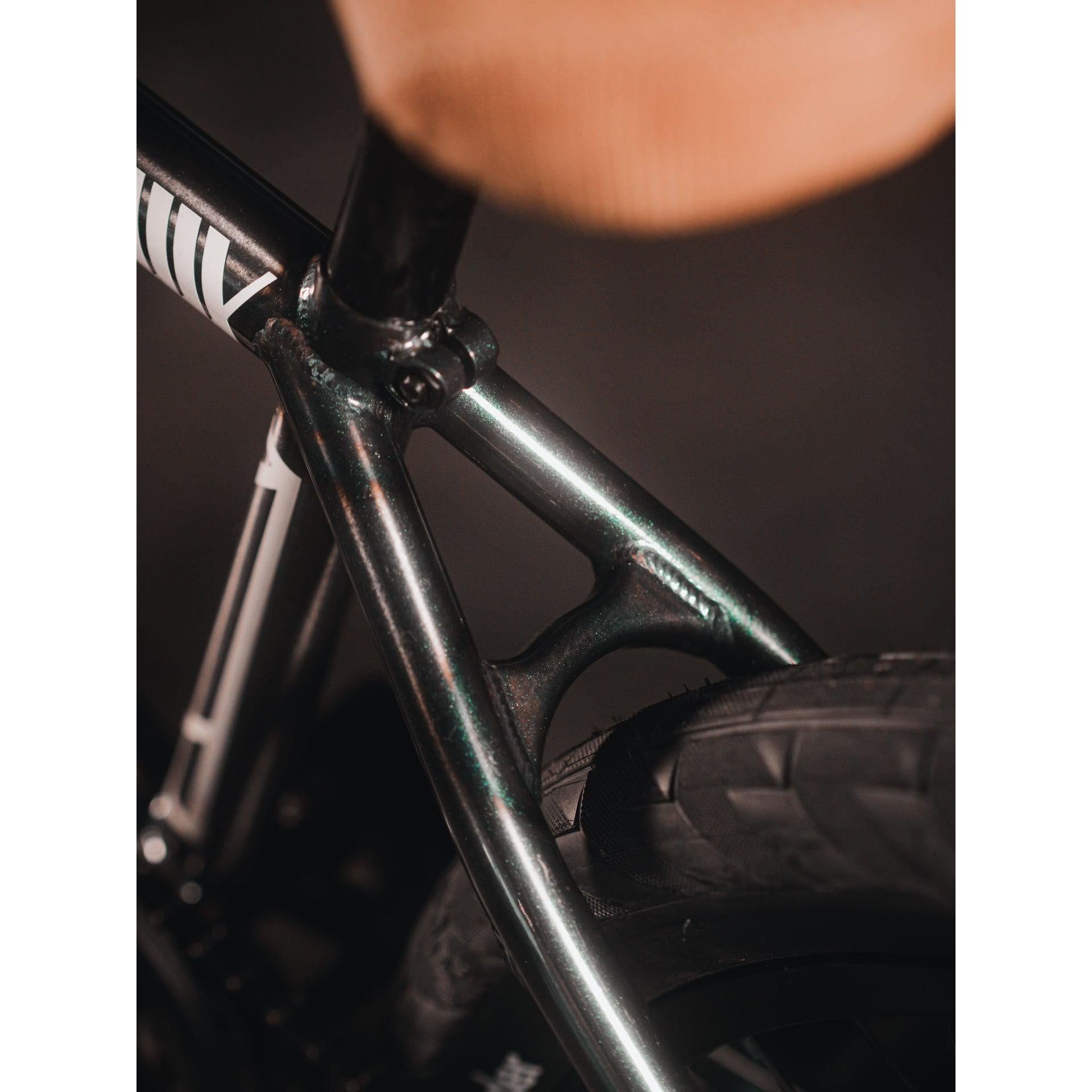 Close-up of a Kink Tactic Custom 20 Inch Bike frame and rear wheel. The image highlights the connection point of the saddle, the upper tubes, and the top of the rear wheel, showcasing a high-quality custom bike. The scene has dim lighting.