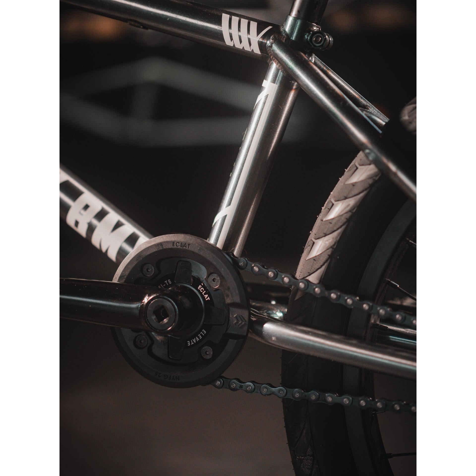 Close-up of a Kink Tactic Custom 20 Inch Bike's chainring, crankset, and rear wheel. The bike frame features metallic elements with white decals.