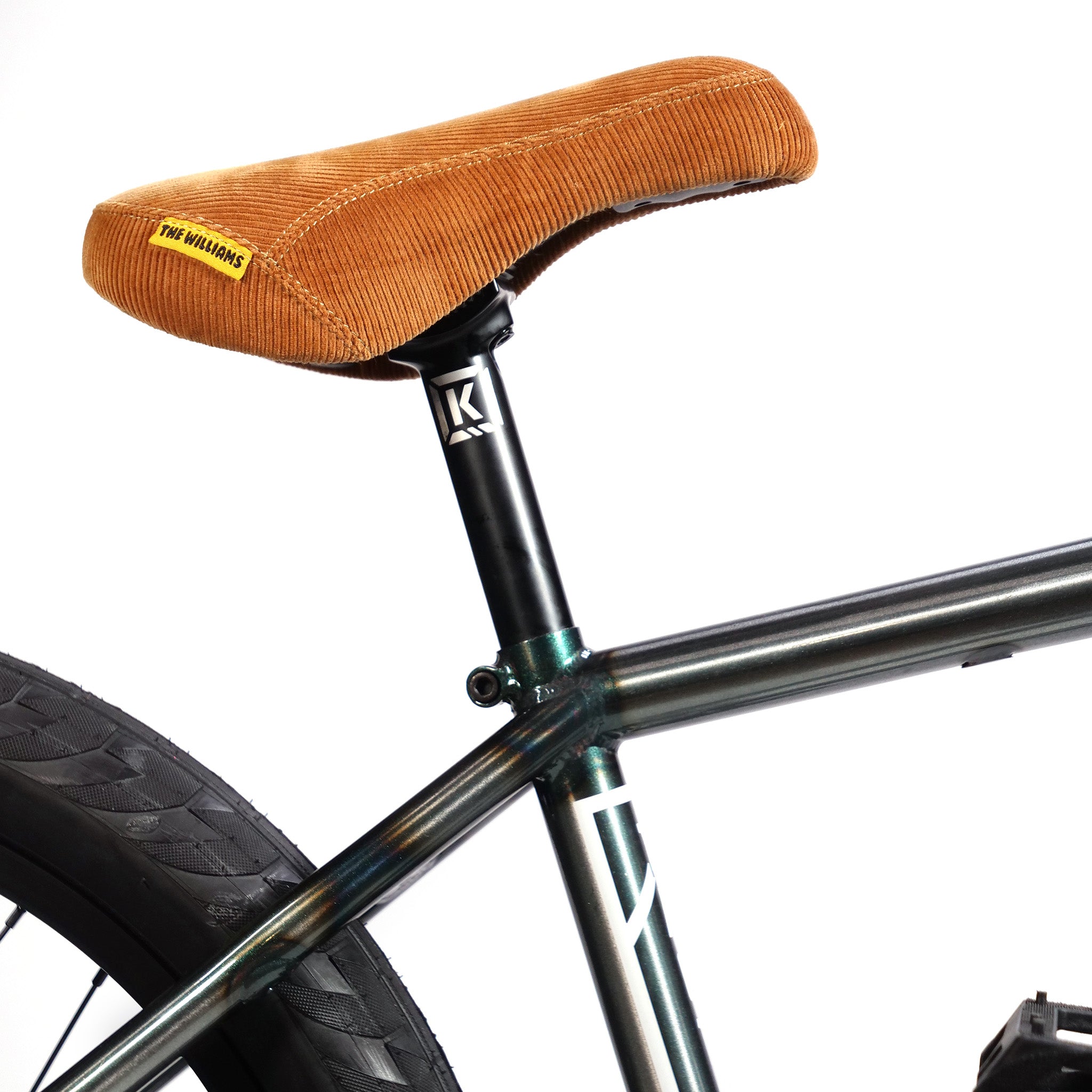 Close-up of a bicycle seat with a brown corduroy cover and a yellow tag, mounted on a black seat post attached to a Kink Tactic frame. The rear wheel is partially visible on this budget-friendly Kink Tactic Custom 20 Inch Bike from LUXBMX.