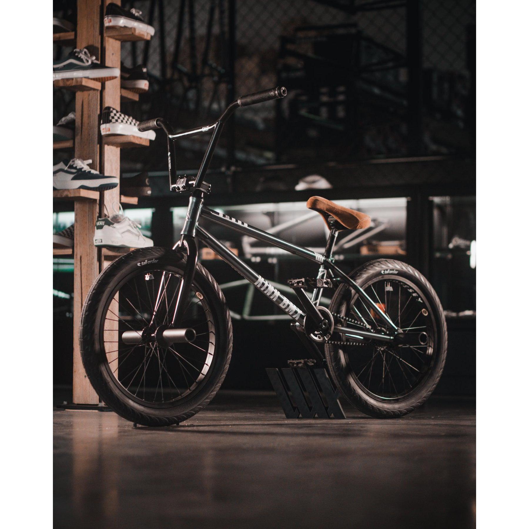 A Kink Tactic Custom 20 Inch Bike with a brown seat is displayed indoors near a wooden shoe rack and other sports equipment.