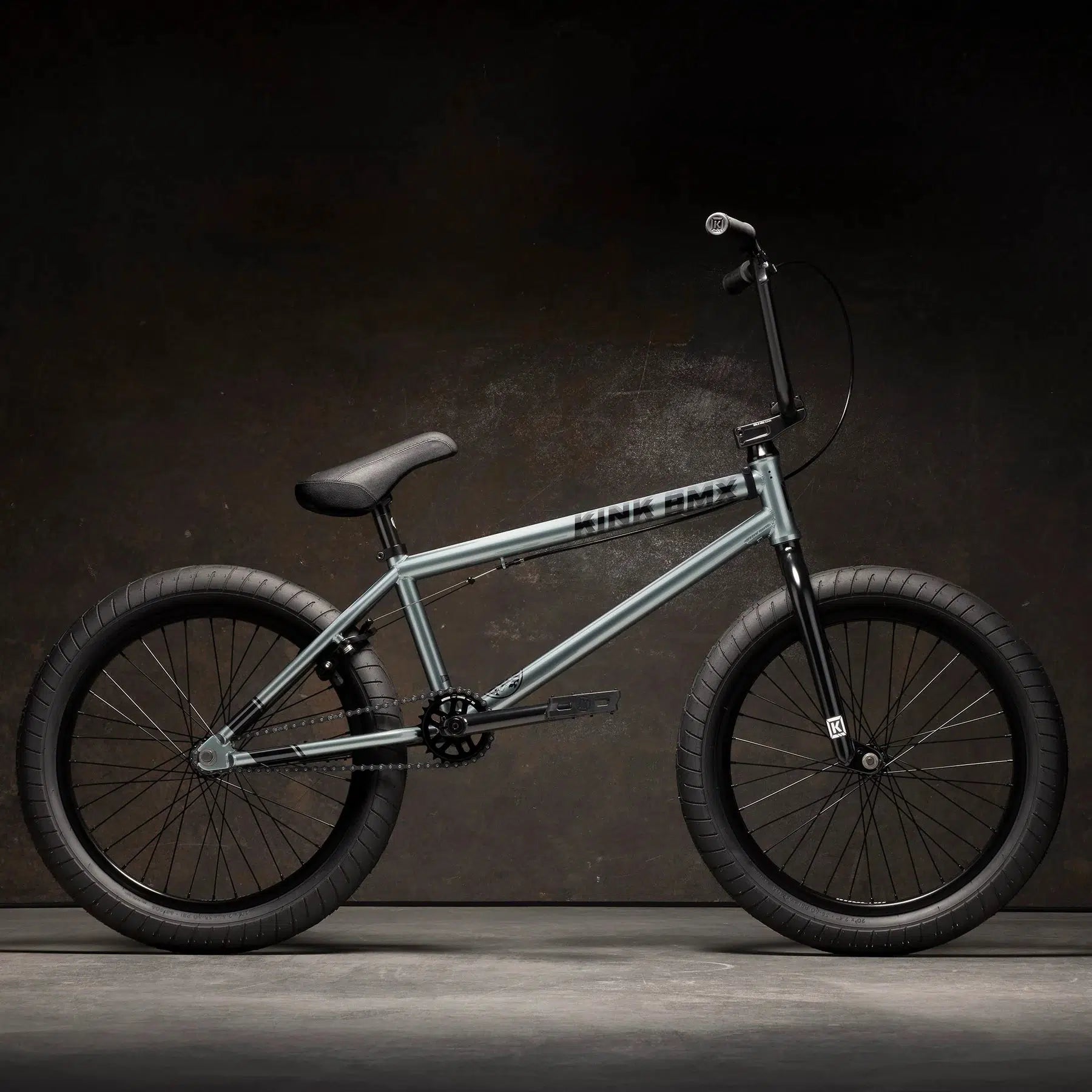 Discover the Kink Whip 20 Inch Bike, a sleek BMX with a gray Chromoly frame, paired perfectly with black handlebars, saddle, and wheels, all set against a dark backdrop.