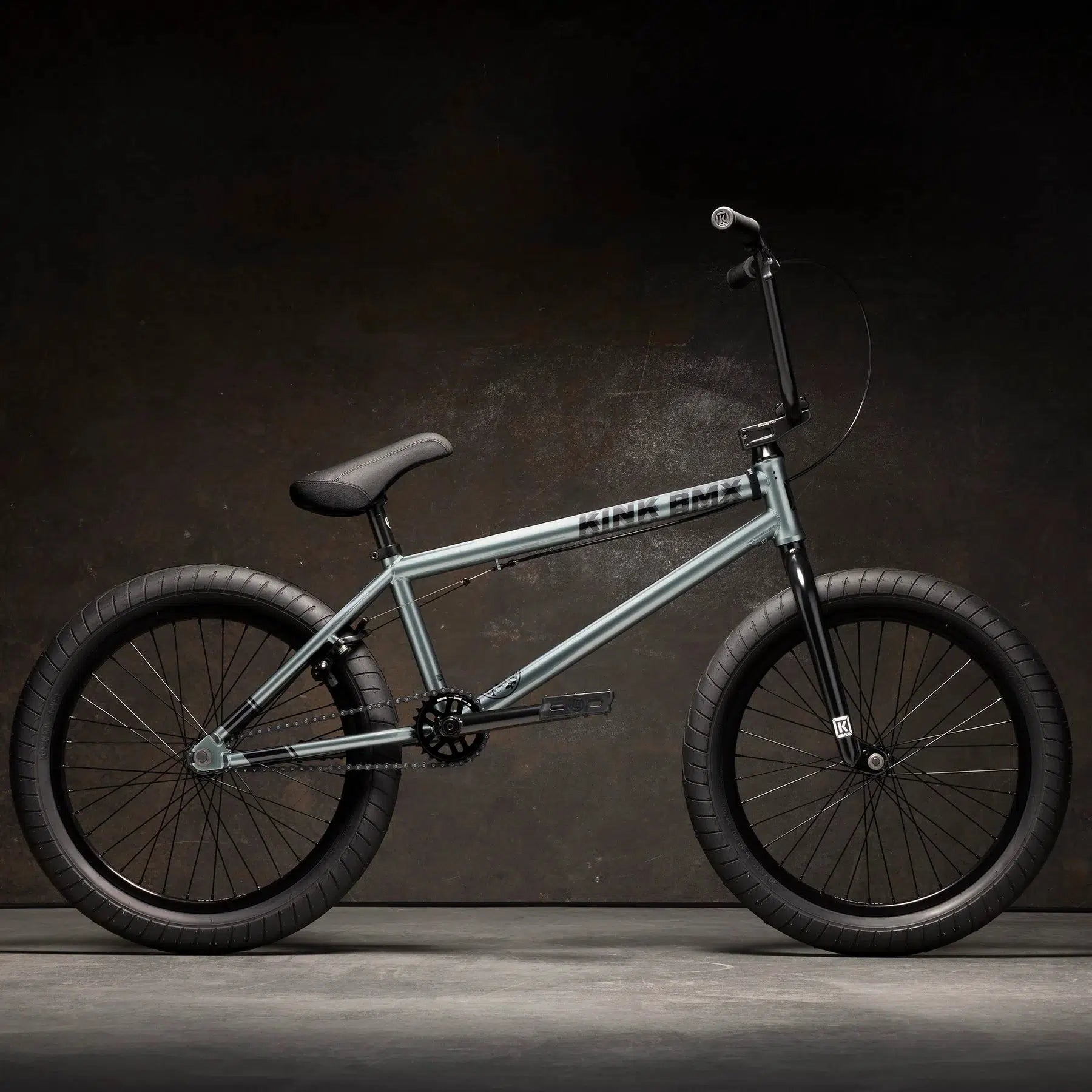 The Kink Whip XL 20 Inch Bike, featuring a robust Chromoly frame in silver with black handlebars and tires, is displayed against a dark background.