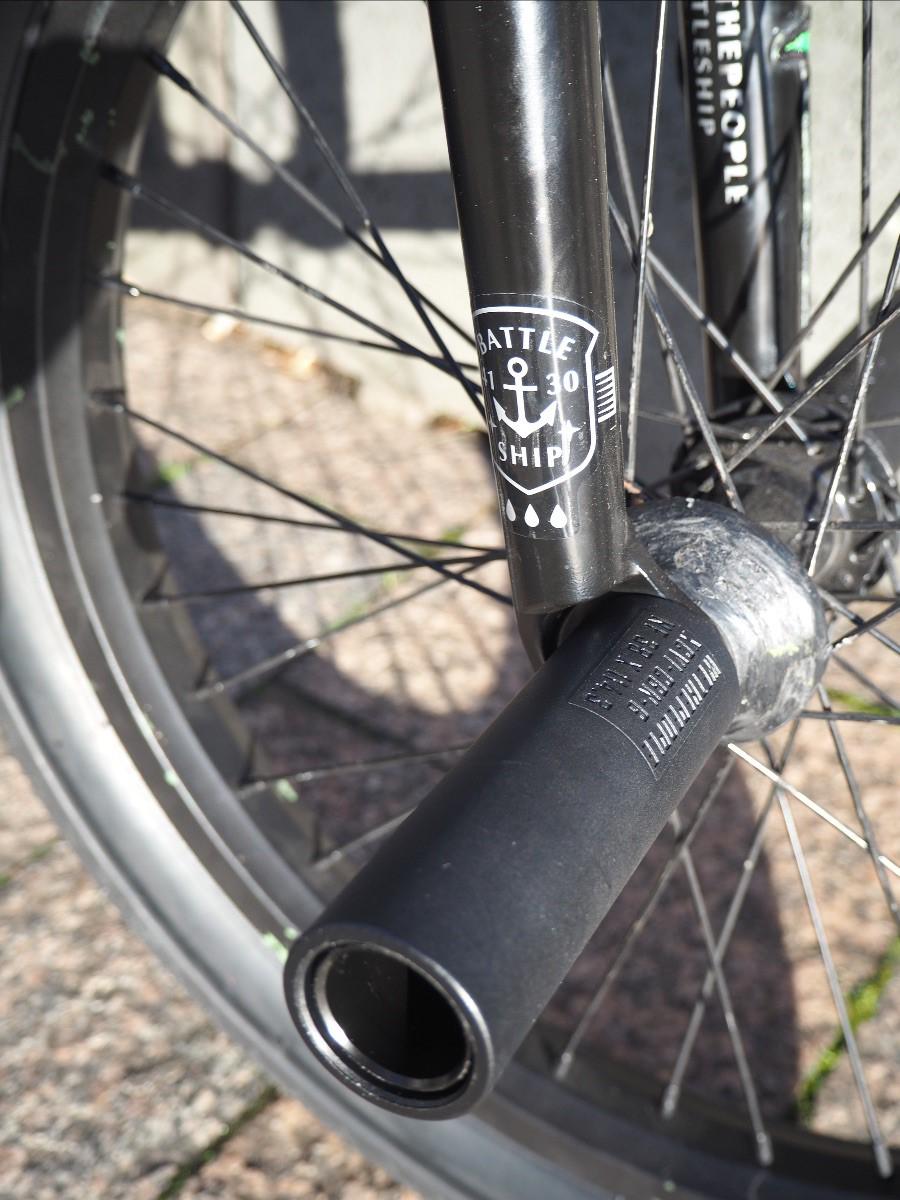 Close-up of a BMX bike front wheel with black spokes, a fork labeled "BATTLESHIP," and a pair of Wethepeople Temper Pegs, featuring a durable chromoly core, set against pavement and a concrete curb.