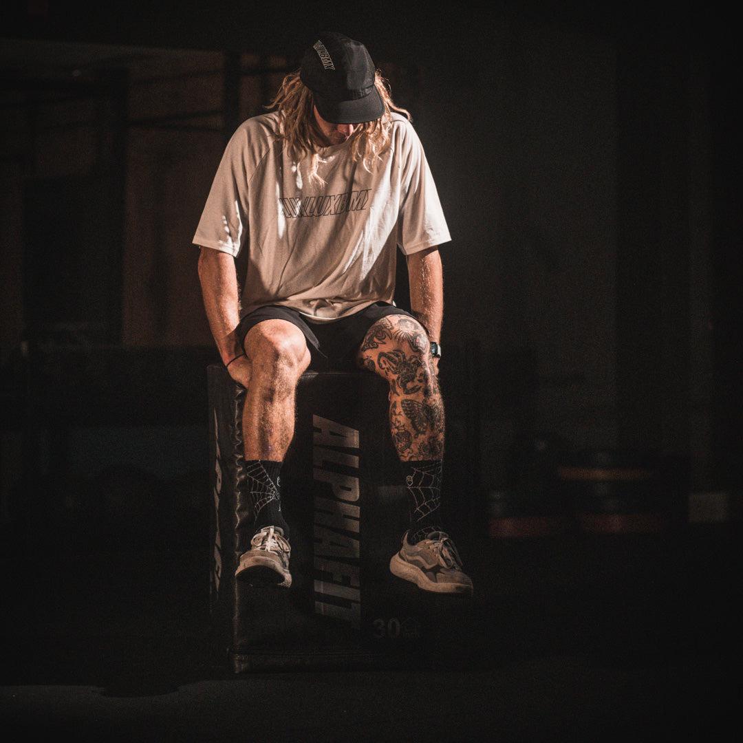 A man, wearing the LUXBMX Bike Athletics T-Shirt - Ecru, sits on a box in a gym.