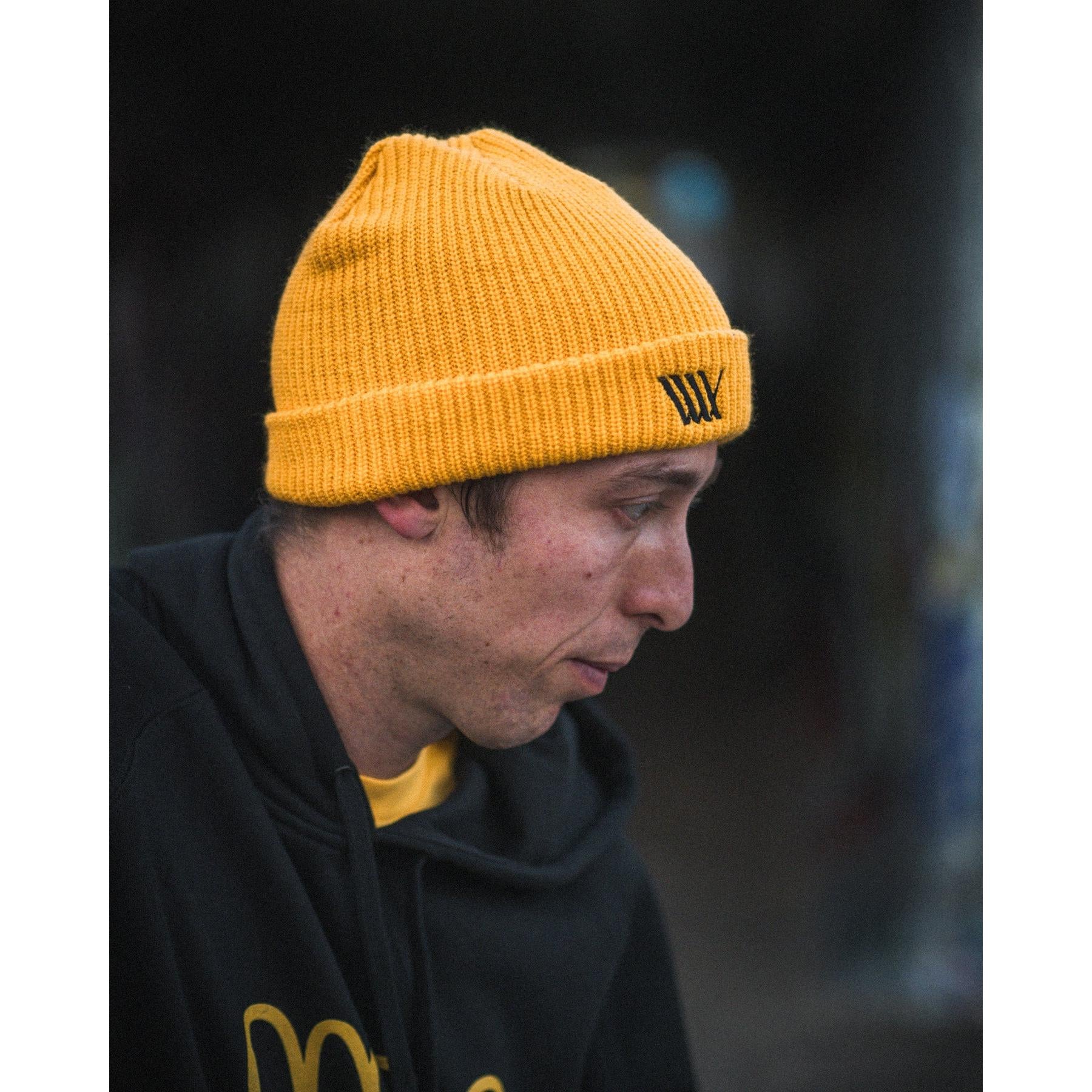 Wearing a yellow LUXBMX Flag Beanie and a black hoodie, a person is shown in profile against a dark, blurred background.