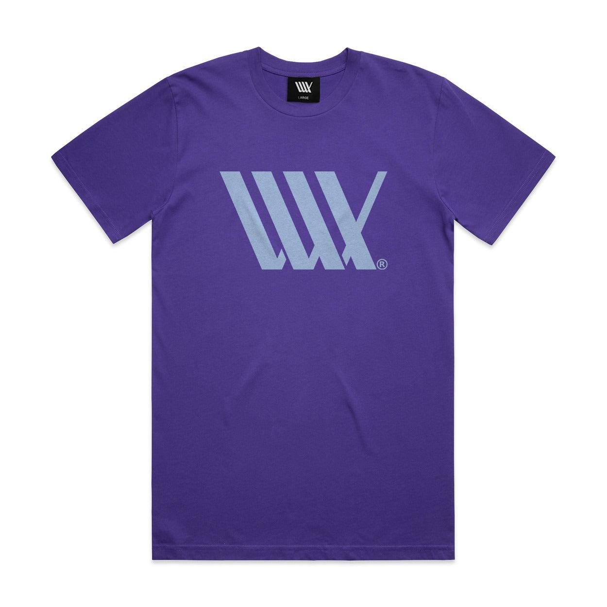 The LUXBMX Flag Tee Violet is a heavyweight, violet T-shirt featuring a light blue geometric logo on the chest. Crafted from combed cotton for superior comfort, it includes a black neckline tag with a white logo. The T-shirt is displayed on a clean white background.