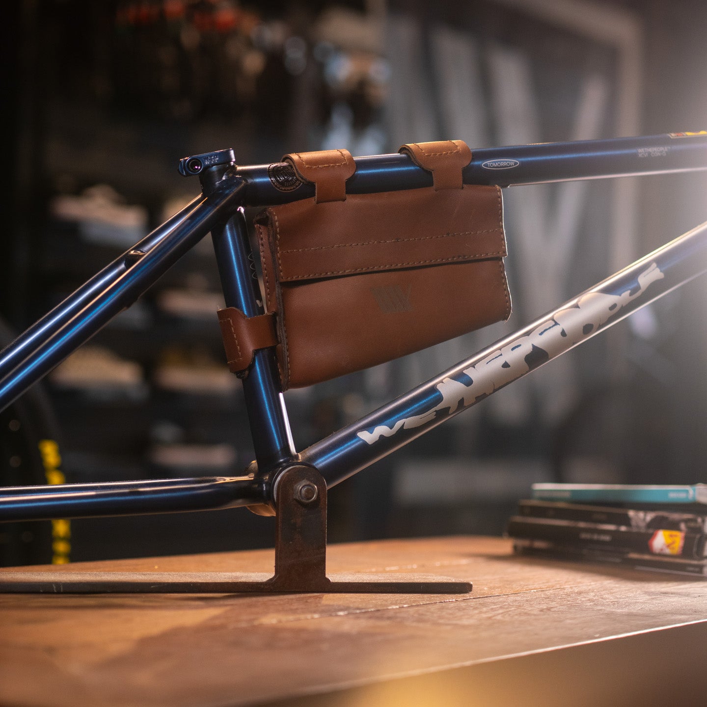 A close-up of a blue bike frame featuring the LUXBMX Leather Frame Bag, shown indoors on a wooden surface with tools and parts in the background, highlights this stylish handmade leather bag as an ideal cycling accessory.