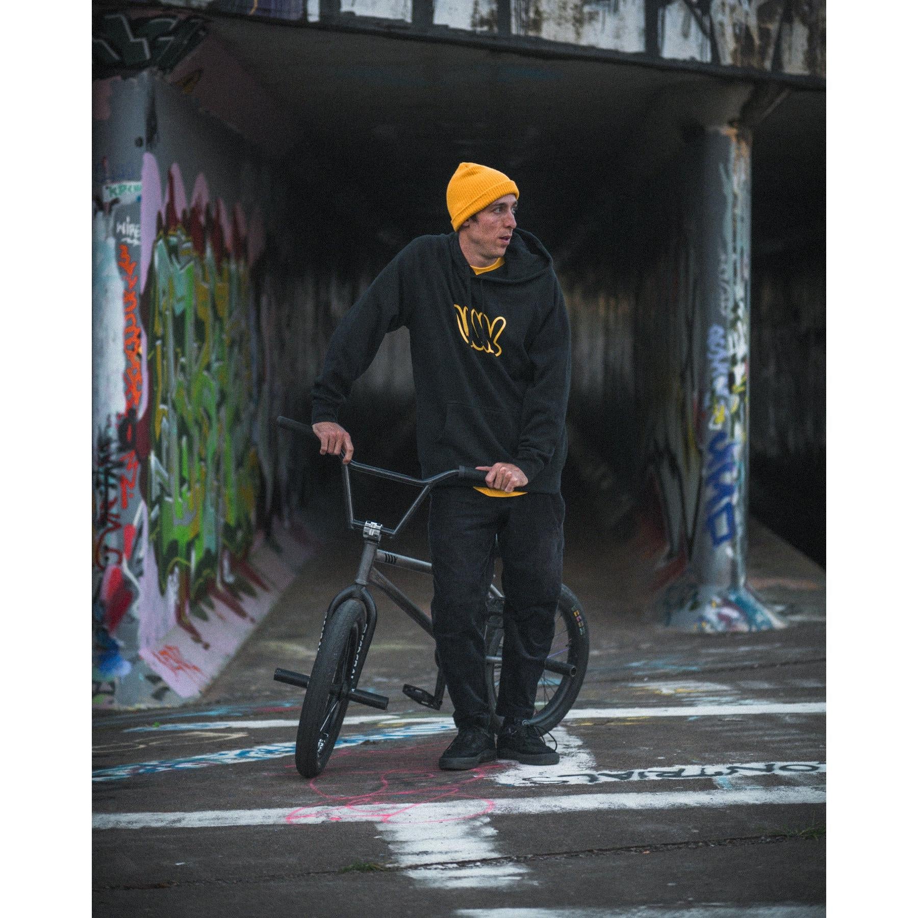 A person in a yellow beanie and the LUXBMX Throwie Hoodie - Black stands beside a BMX bike on a graffiti-covered path under a bridge.