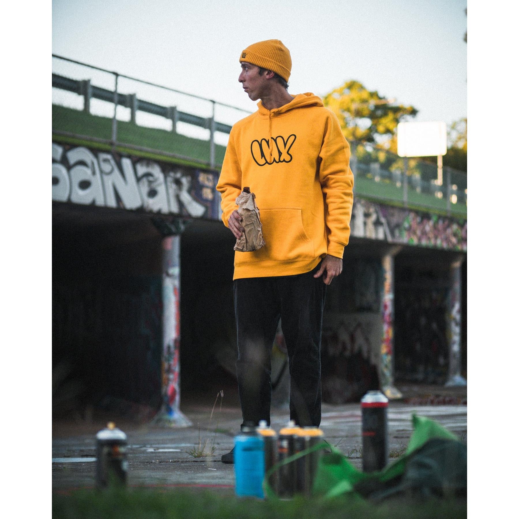 Wearing a yellow LUXBMX Throwie Hoodie, a person stands outdoors by a graffiti wall, holding a paper bag with spray paint cans at their feet.