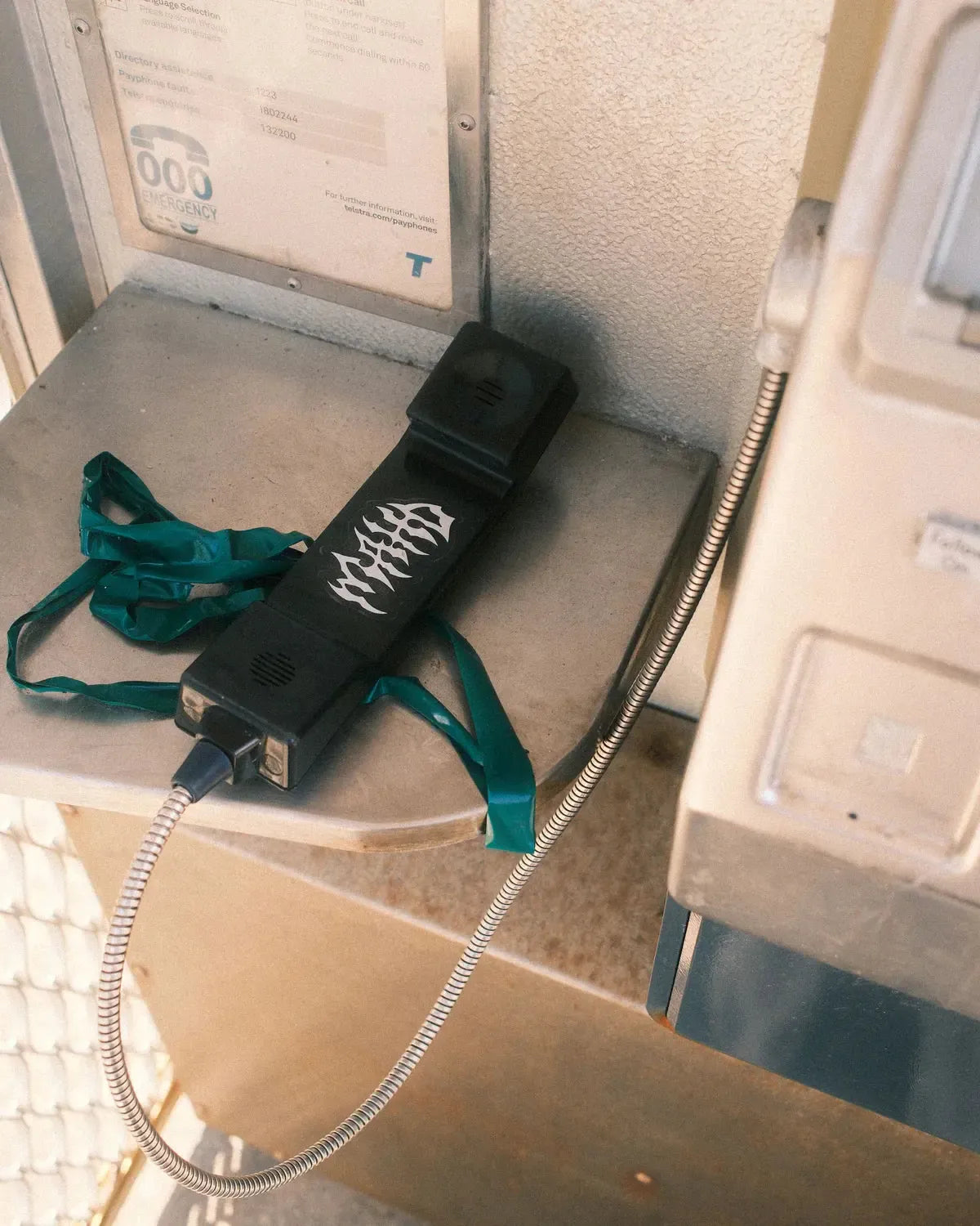 A Mahd Metal Tee in sand color with a green ribbon is draped off the hook, covered in graffiti, next to a payphone displaying a subtle Centre logo.