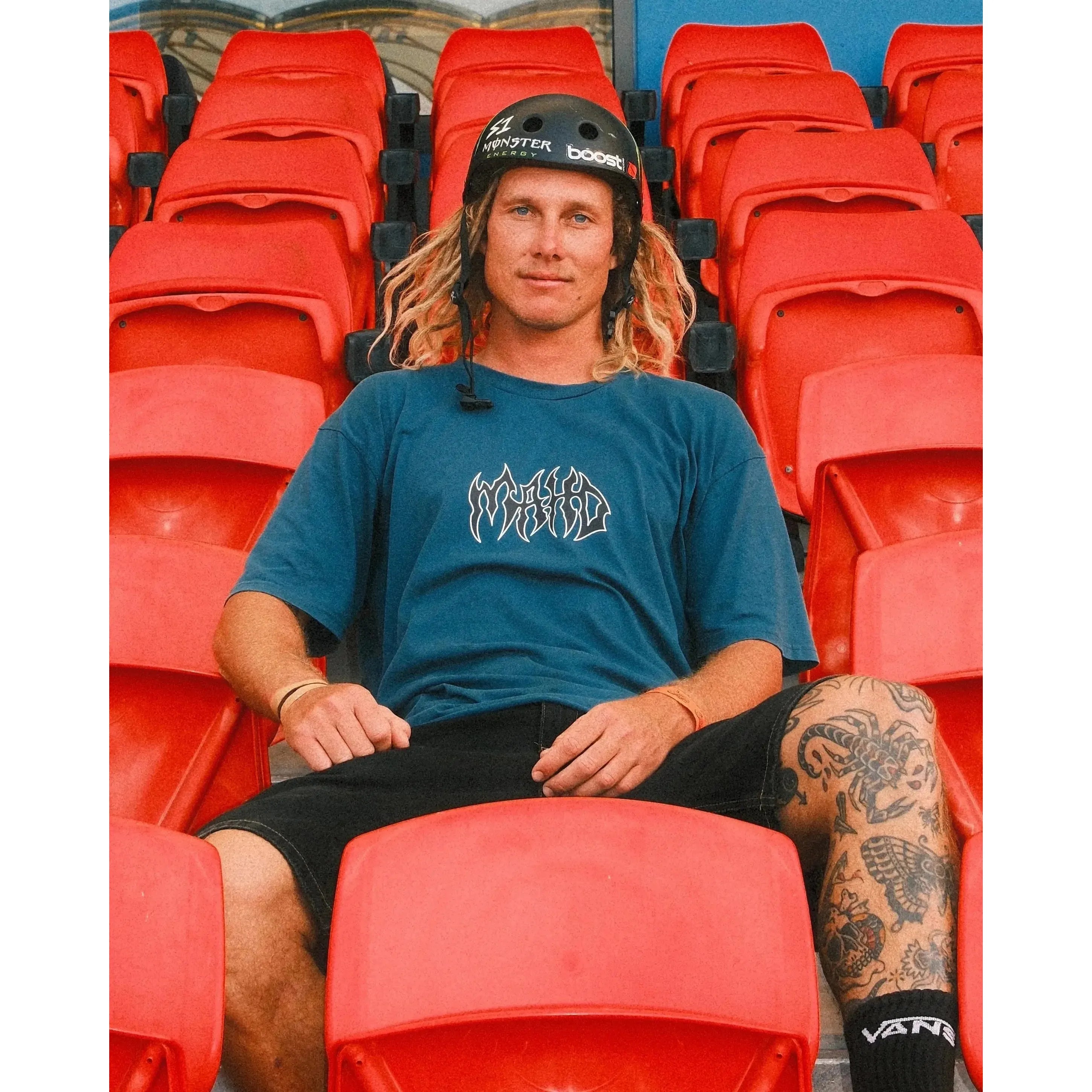 A person with a helmet and dreadlocks sits on red stadium seats, wearing the blue Mahd Metal Tee with a centre logo, black shorts, and showcasing a leg tattoo.