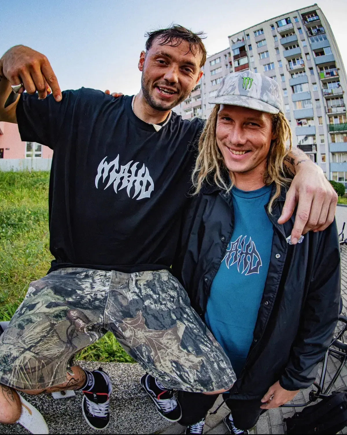 Two men pose outside near apartments; one points to his black Mahd Metal Tee with a bold center logo, while the other, in a gray cap and blue graphic tee with a central design, stands beside him.
