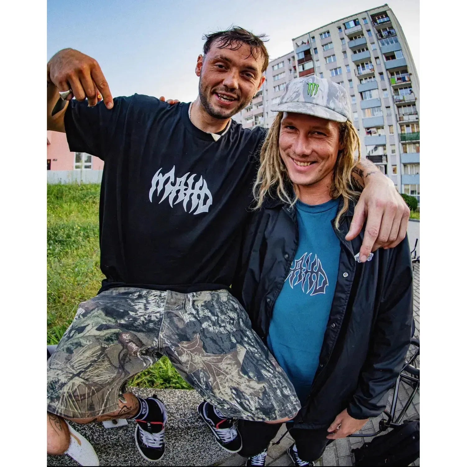 Two men pose outside near apartments; one points to his black Mahd Metal Tee with a bold center logo, while the other, in a gray cap and blue graphic tee with a central design, stands beside him.