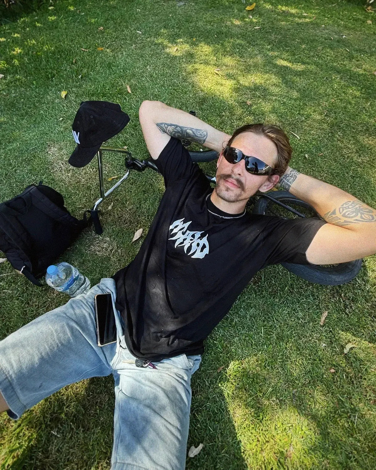 A man wearing the sand-coloured Mahd Metal Tee with a centre logo and sunglasses lies on the grass, resting his head on a bicycle wheel. Nearby are a cap, backpack, water bottle, and phone.