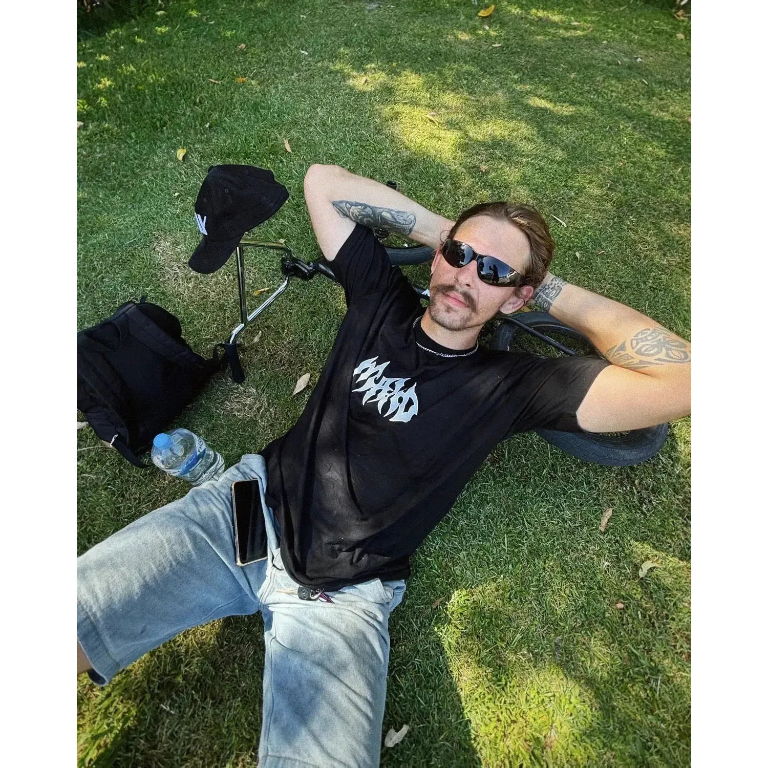 A man wearing the sand-coloured Mahd Metal Tee with a centre logo and sunglasses lies on the grass, resting his head on a bicycle wheel. Nearby are a cap, backpack, water bottle, and phone.