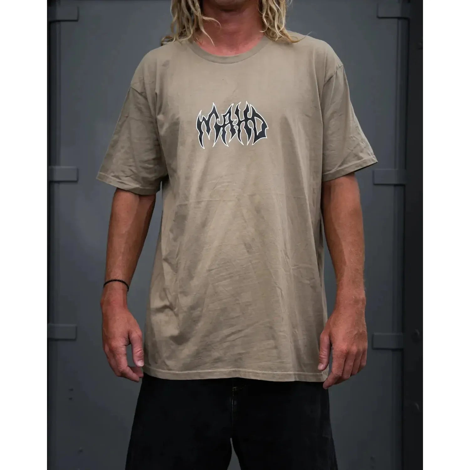 A person wears a sand Mahd Metal Tee featuring a central "World World" logo, standing in front of a gray metal background.