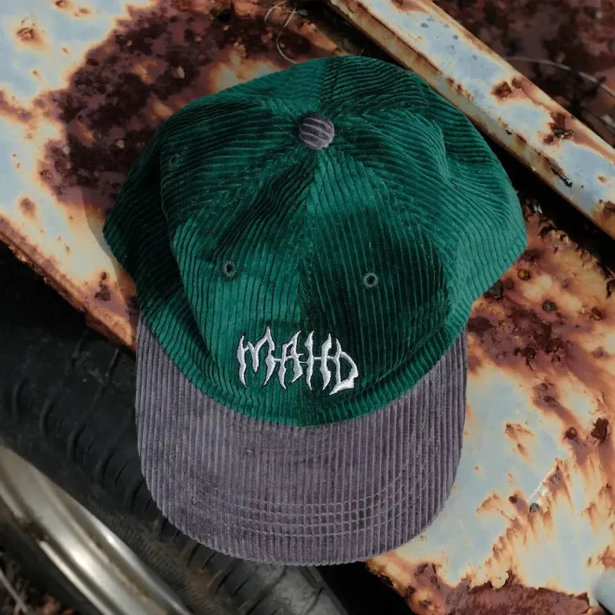 A green and gray Mahd Metal Corduroy Hat with "MAID" embroidered on the crown, displayed on a rusty metal surface.