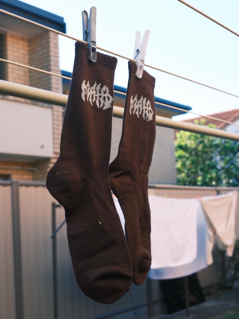 A pair of Mahd Metal Socks, brown with white designs, hang from a clothesline by clothespins. Made from a bamboo blend, these one-size-fits-most socks offer everyday comfort against a backdrop of laundry in a suburban backyard.
