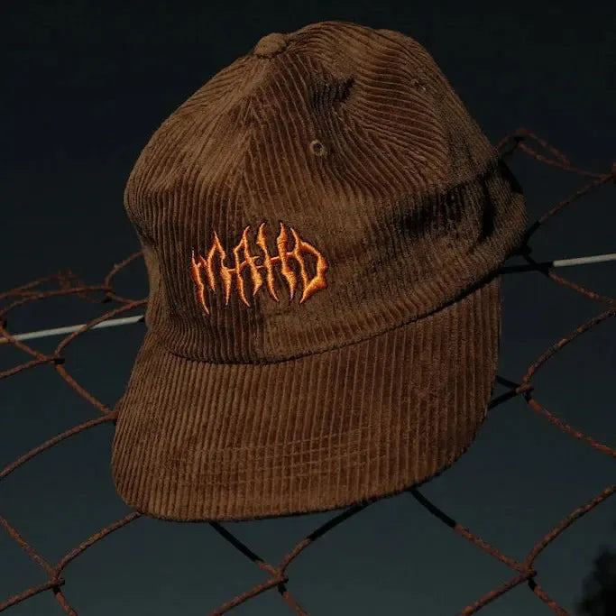 A brown Mahd Metal Corduroy Hat with orange embroidered text rests on a wire fence against a dark background.