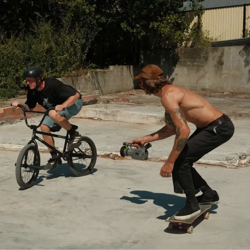 A person, possibly Boyd Hilder in a Mahd Metal Corduroy Hat, rides a BMX bike on concrete while a shirtless skateboarder films with a handheld camera outdoors.