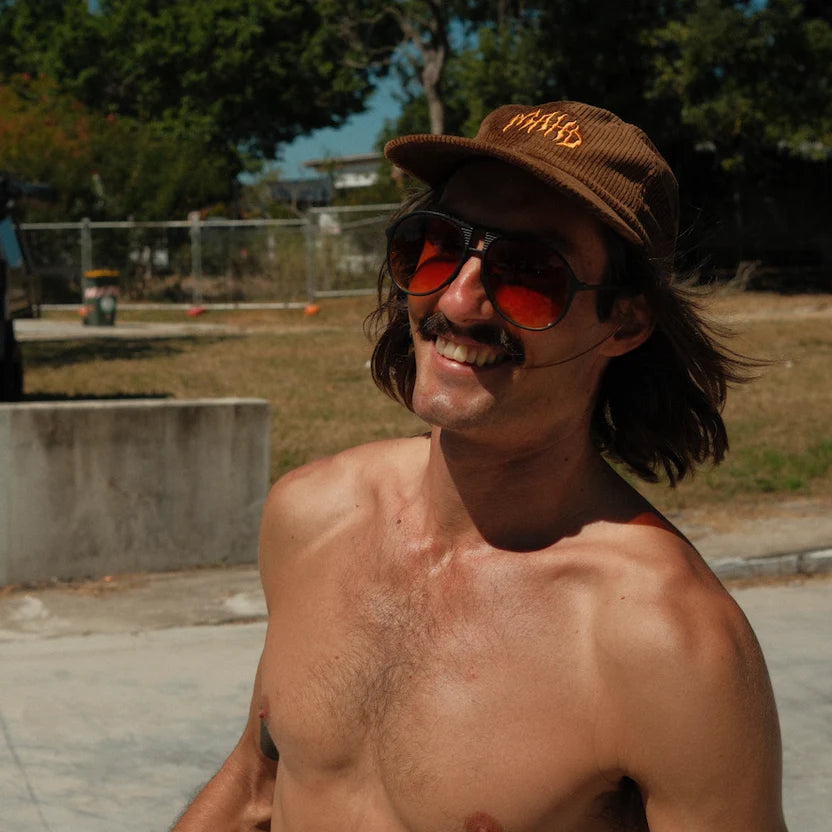 A shirtless man wearing the Mahd Metal Corduroy Hat, large sunglasses, and a mustache smiles outdoors on a sunny day.