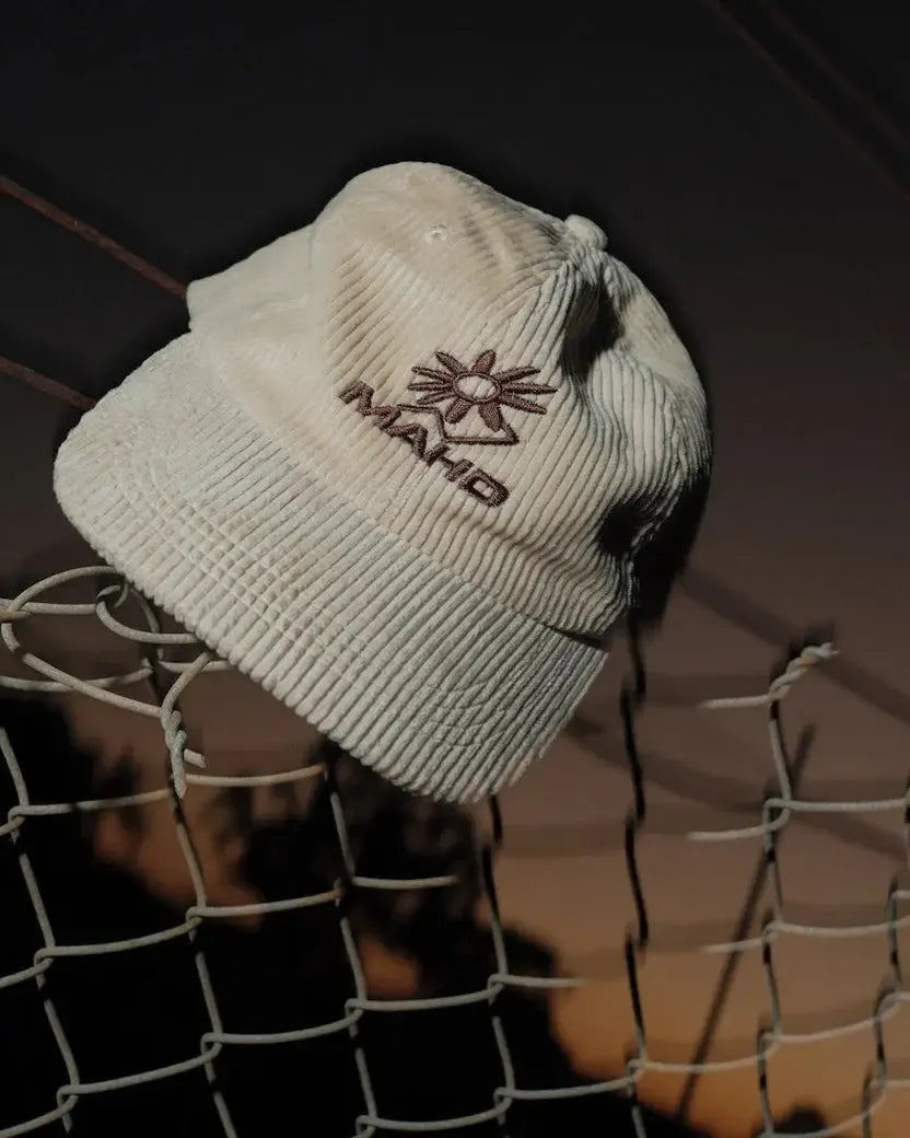 The Mahd Botanical Corduroy Hat, featuring cords and an embroidered "MANTO" logo, sits on a wire fence at dusk, delivering Boyd Hilder-inspired style.