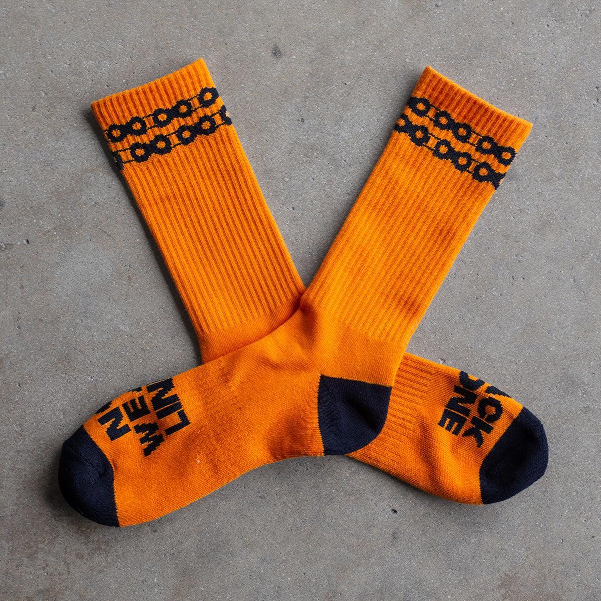 Backbone No Weak Links Socks: Orange socks with black toe and heel, chain link design near the cuffs, and "NO WEAK LINKS" in black on the soles, shown on a concrete surface.