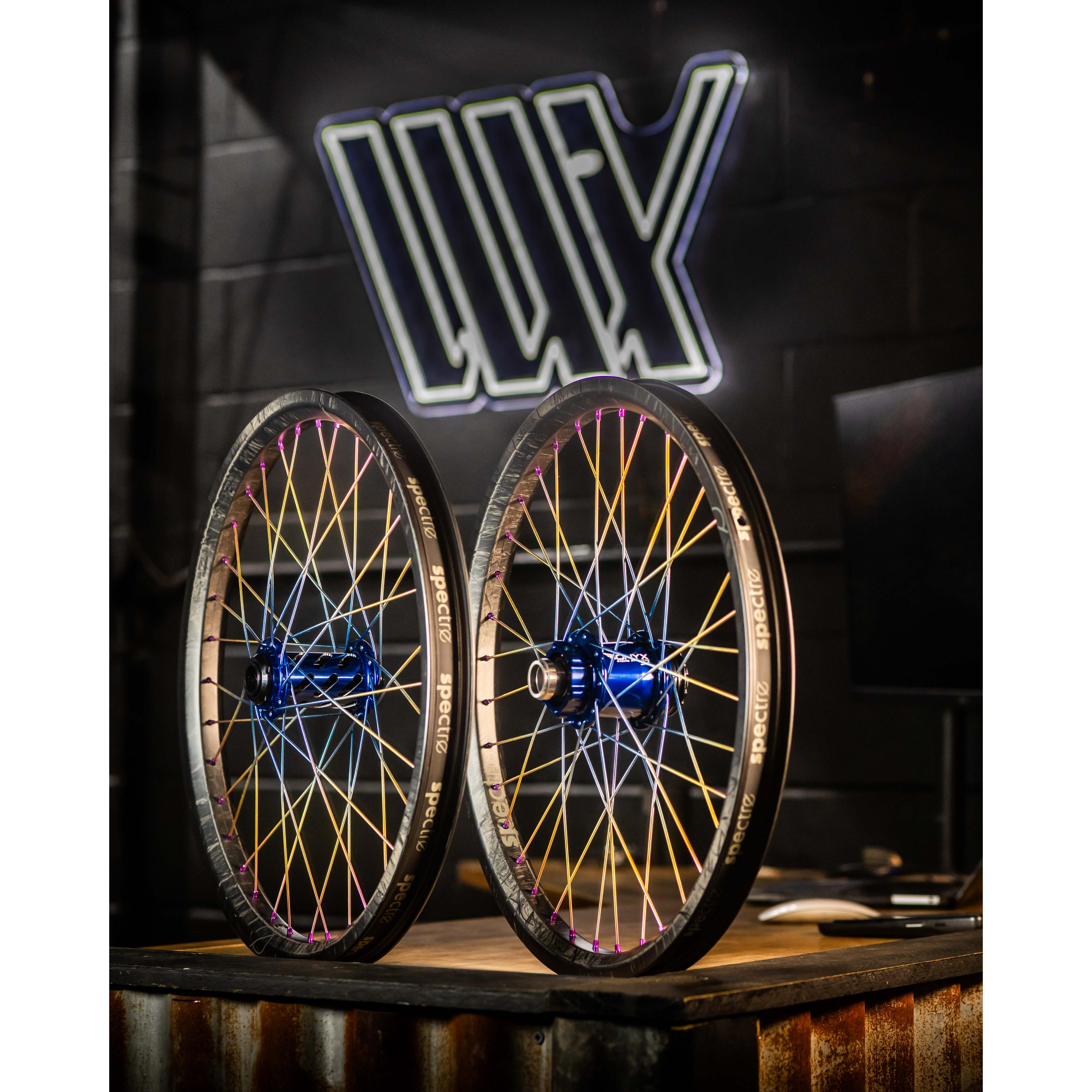 Two Onyx X Spectre Carbon Marble Sunset 20 Inch Wheels with colorful titanium spokes and blue hubs are upright on a wooden table against a dark wall featuring a large, illuminated logo.