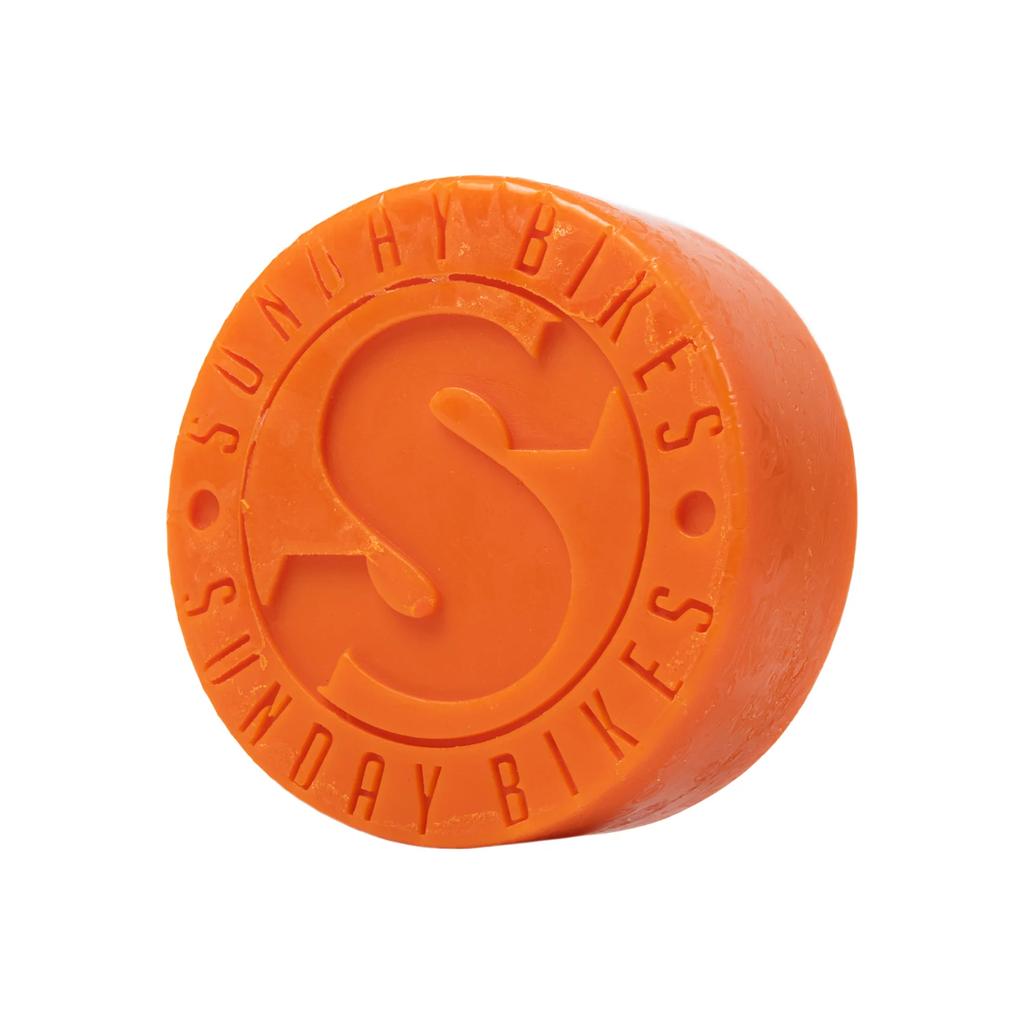 Shaped like an orange puck with "SUNDAY BIKES" and a large "S" embossed, the Sunday Puck Grind Wax is ideal for skateboarders seeking high-quality skateboard wax.