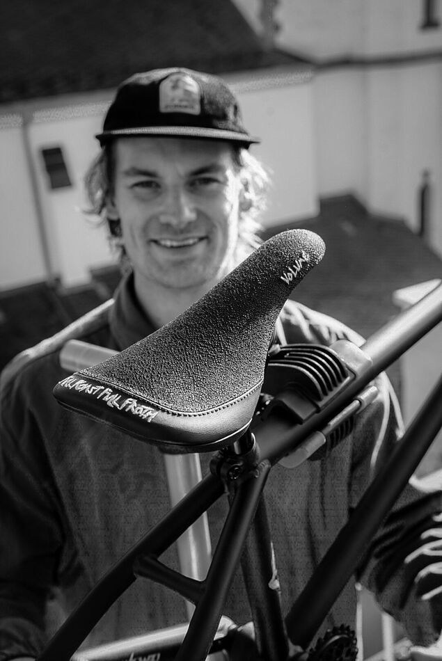 A man in a cap smiles outdoors behind a close-up of the Volume Full Roast Full Froth railed bicycle seat by Paul Theolen, attached to a bike frame.
