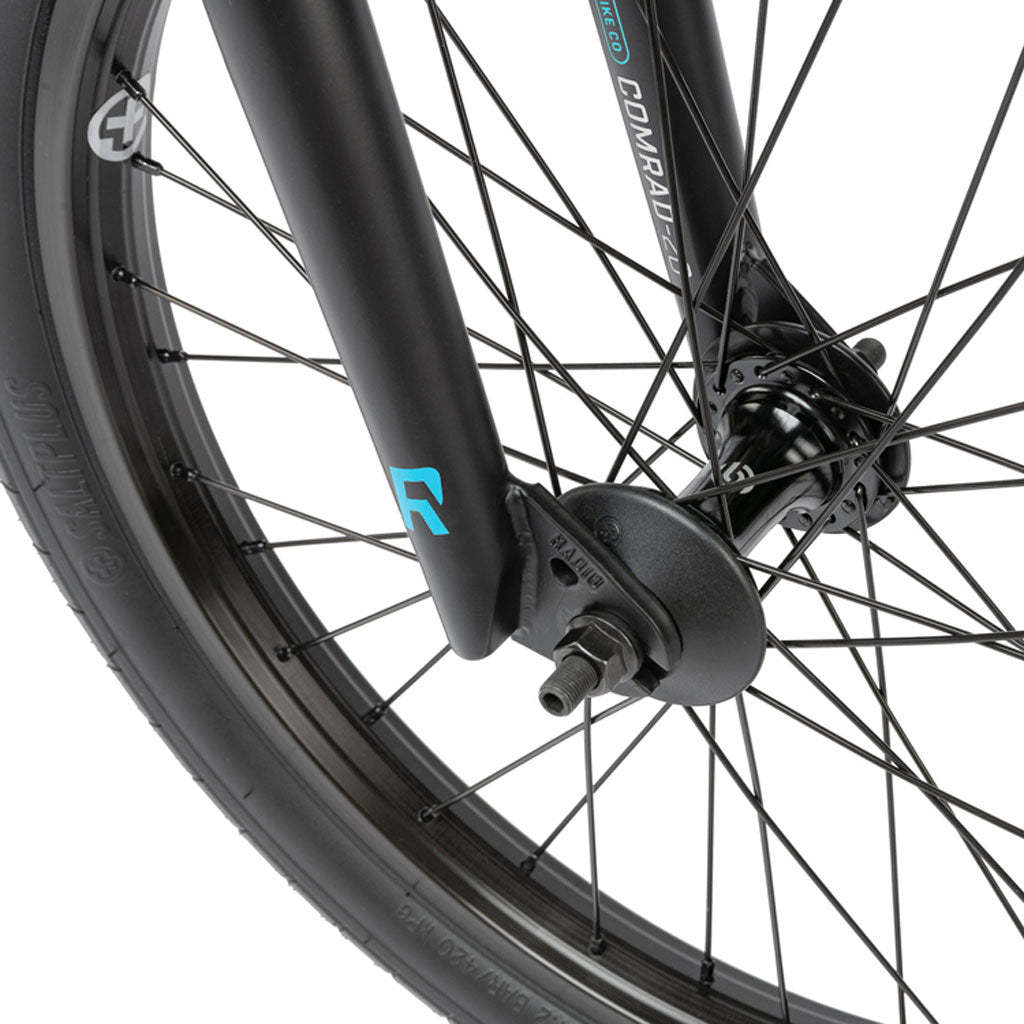 A close-up of the black front wheel with visible spokes and axle highlights the blue and white branding on the fork, perfectly complementing the rugged Radio Comrad 20 inch Bike design.
