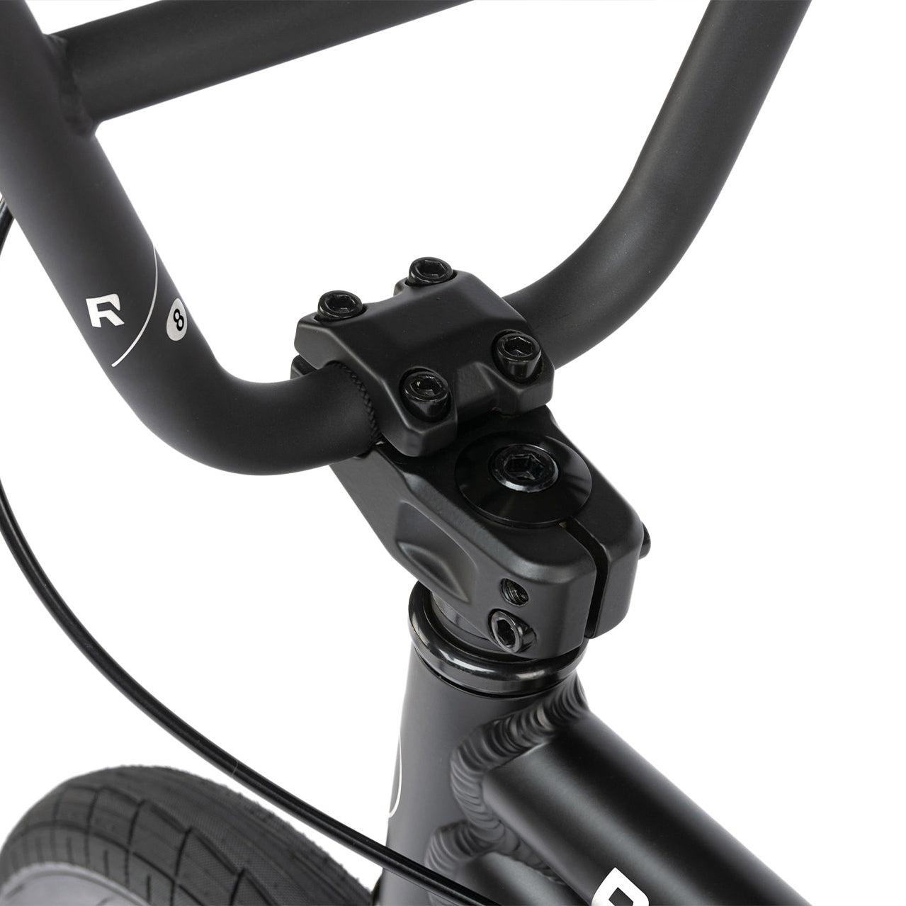 Close-up of a Radio Dice 16 Inch Bike handlebar and front stem, featuring bolts and part of the front wheel. The black handlebar, made from 6061 Alloy, showcases a small logo on the side.