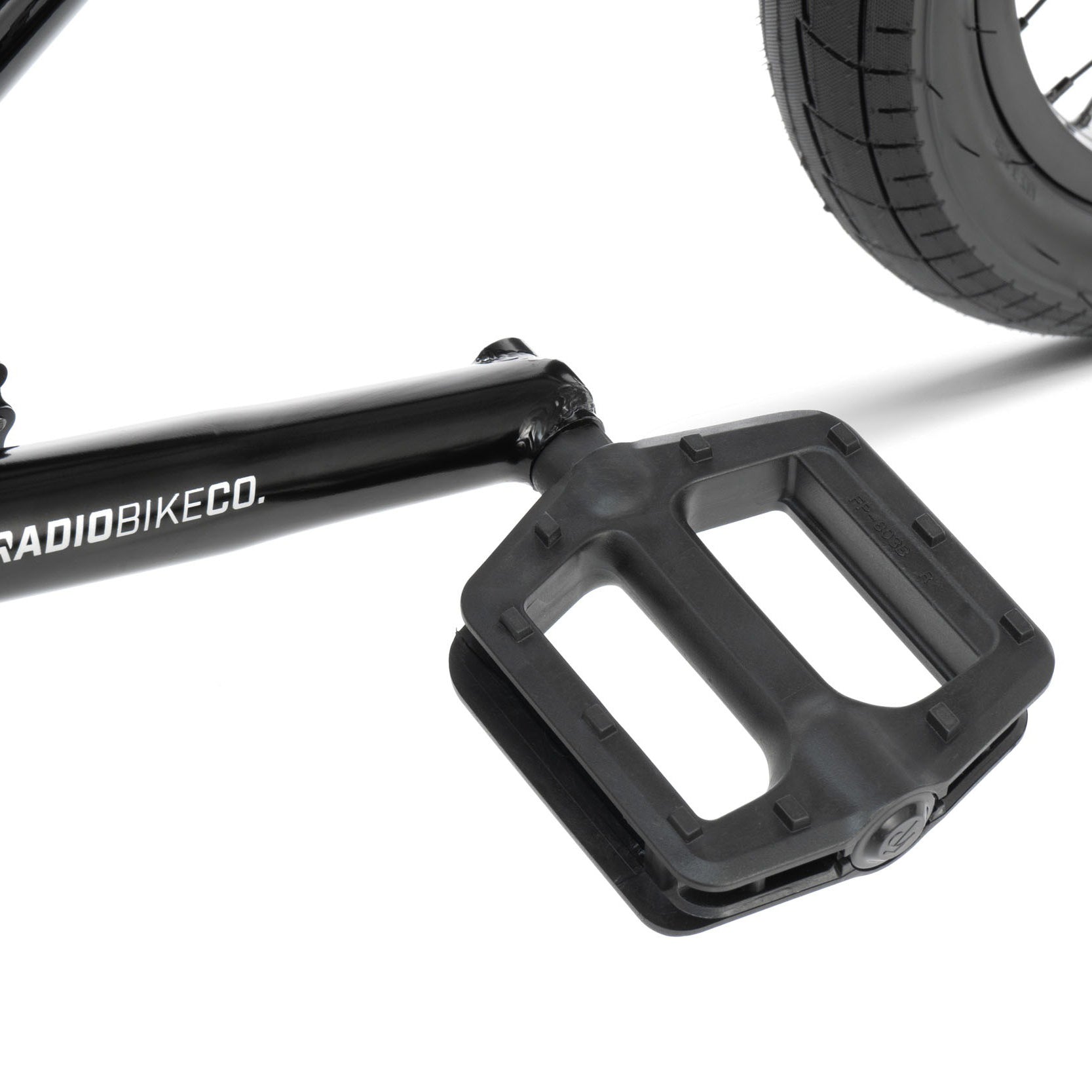 Close-up image of a bicycle pedal and crank arm attached to the frame, featuring Radio Dice 18 Inch Bike with black components and a portion of the rear tire visible.