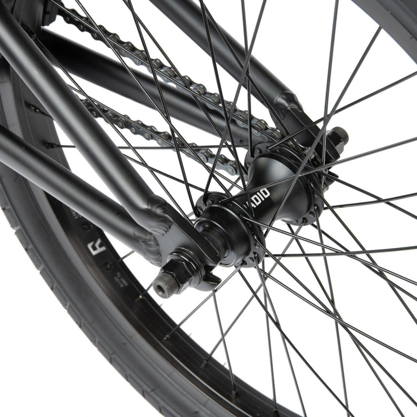 Close-up of a black bicycle wheel with visible spokes, tire, and center hub featuring a chain in the background, showcasing the sturdy Radio Dice 20 Inch Bike.