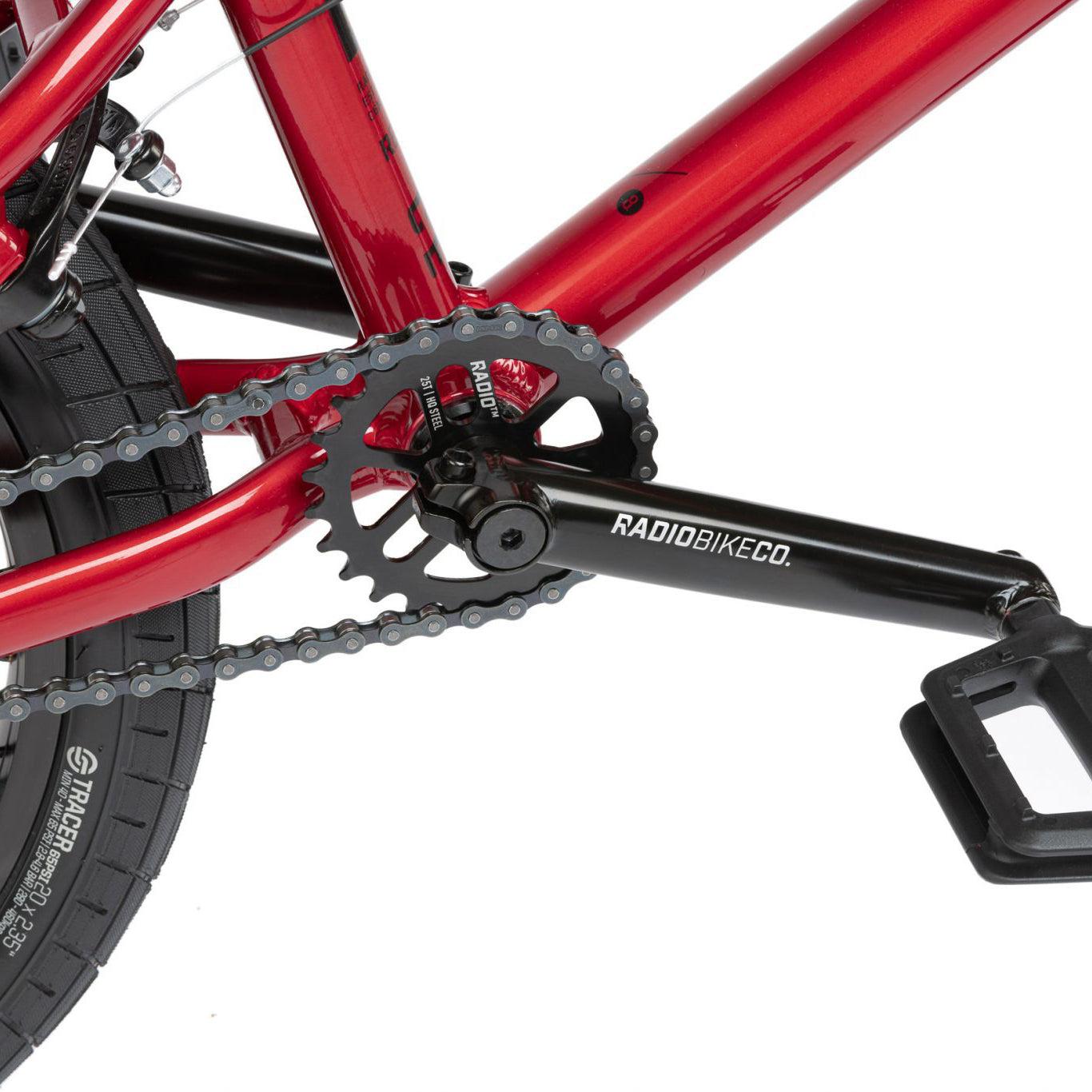 Close-up of a red Radio Dice 20 Inch Bike's crankset, chainring, and pedal. The chain is visible around the chainring. The side of the tire reads "TRACTION" featuring Salt Tracer Tyres, and the crank arm is labeled "RADIOBIKECO.