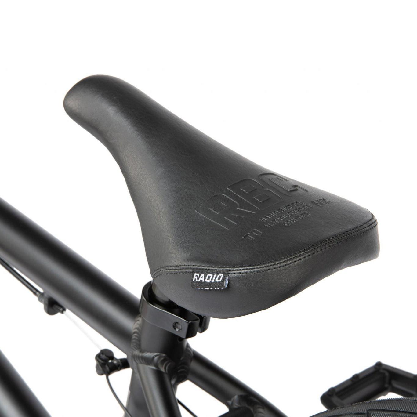 A close-up of a black bicycle saddle with the brand name "RADIO" embossed on it, attached to the seat post of a sleek Radio Dice 20 Inch Bike with a strong 6061 Alloy Frame.