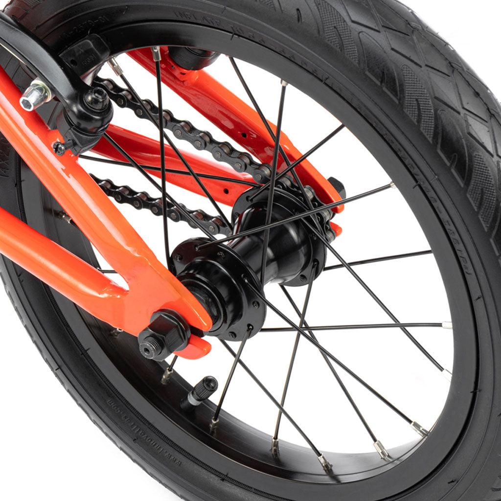 Close-up view of a BMX bike's rear wheel and chain mechanism showcasing a black hub, spokes, tire, and the vibrant orange frame of the Radio Revo 14 Inch bike.