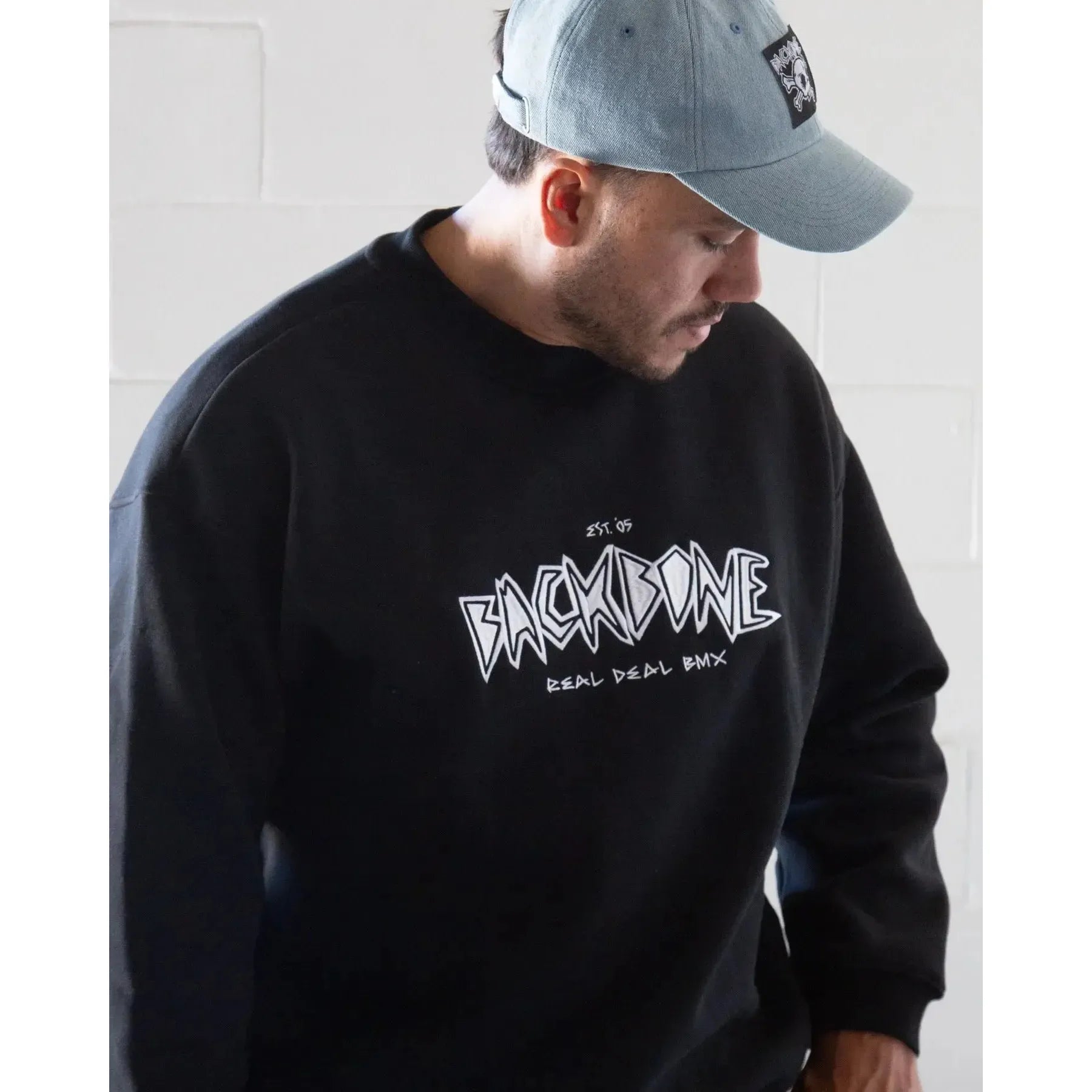 A man in a light blue cap and a Backbone Real Deal Crewneck featuring "BACKBONE REAL DEAL BMX" on the front stands against a white tiled wall.