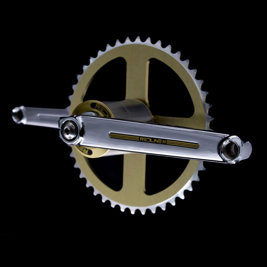 A close-up of the REDLINE Flight 401 50th Anniversary Crank Set in gold and silver, showcasing "Redline" branding and a large chainring, set against a black background.