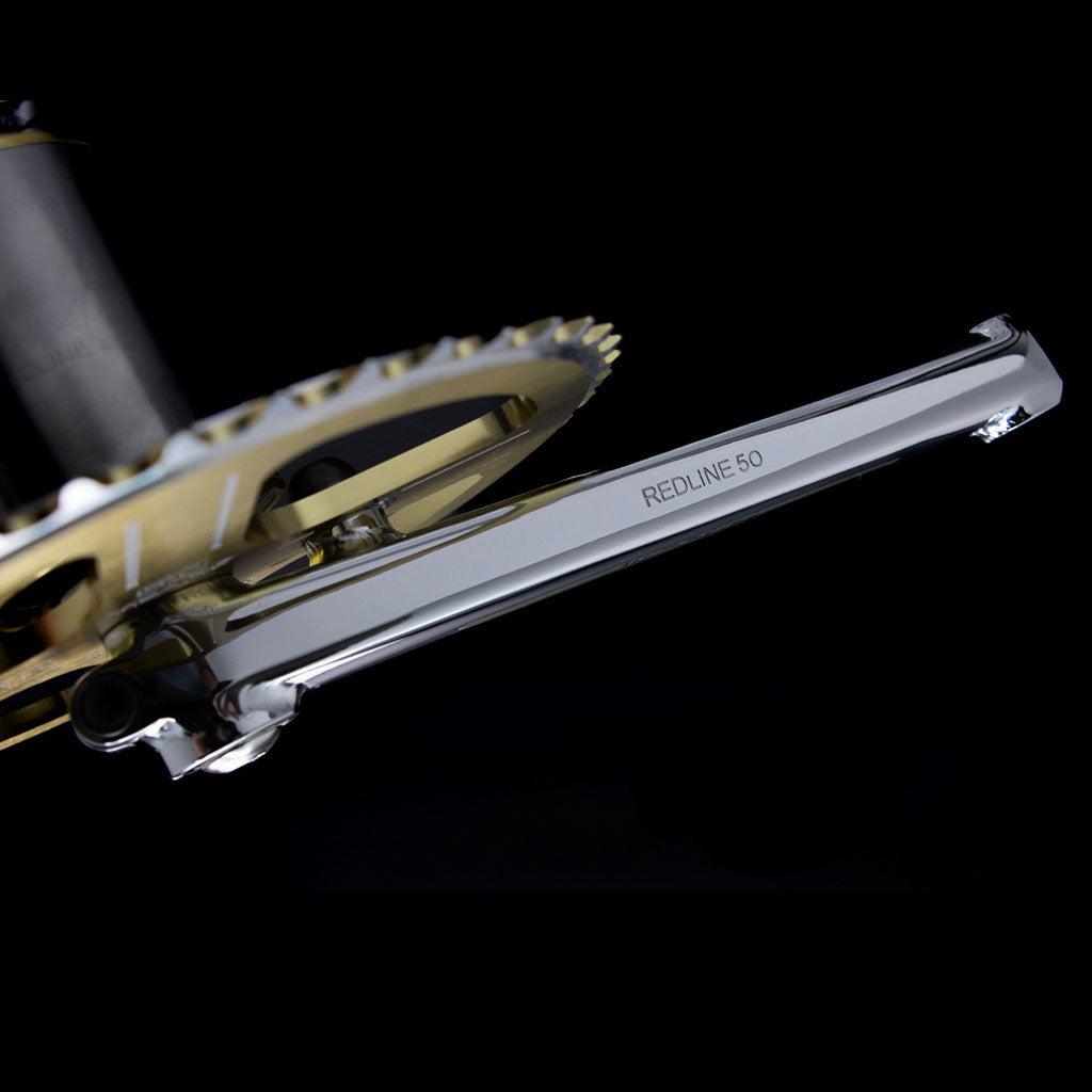 Close-up of a REDLINE Flight 401 50th Anniversary Crank Set labeled "REDLINE 50," attached to a gold chainring, commemorating 50 years of BMX, set against a black background.